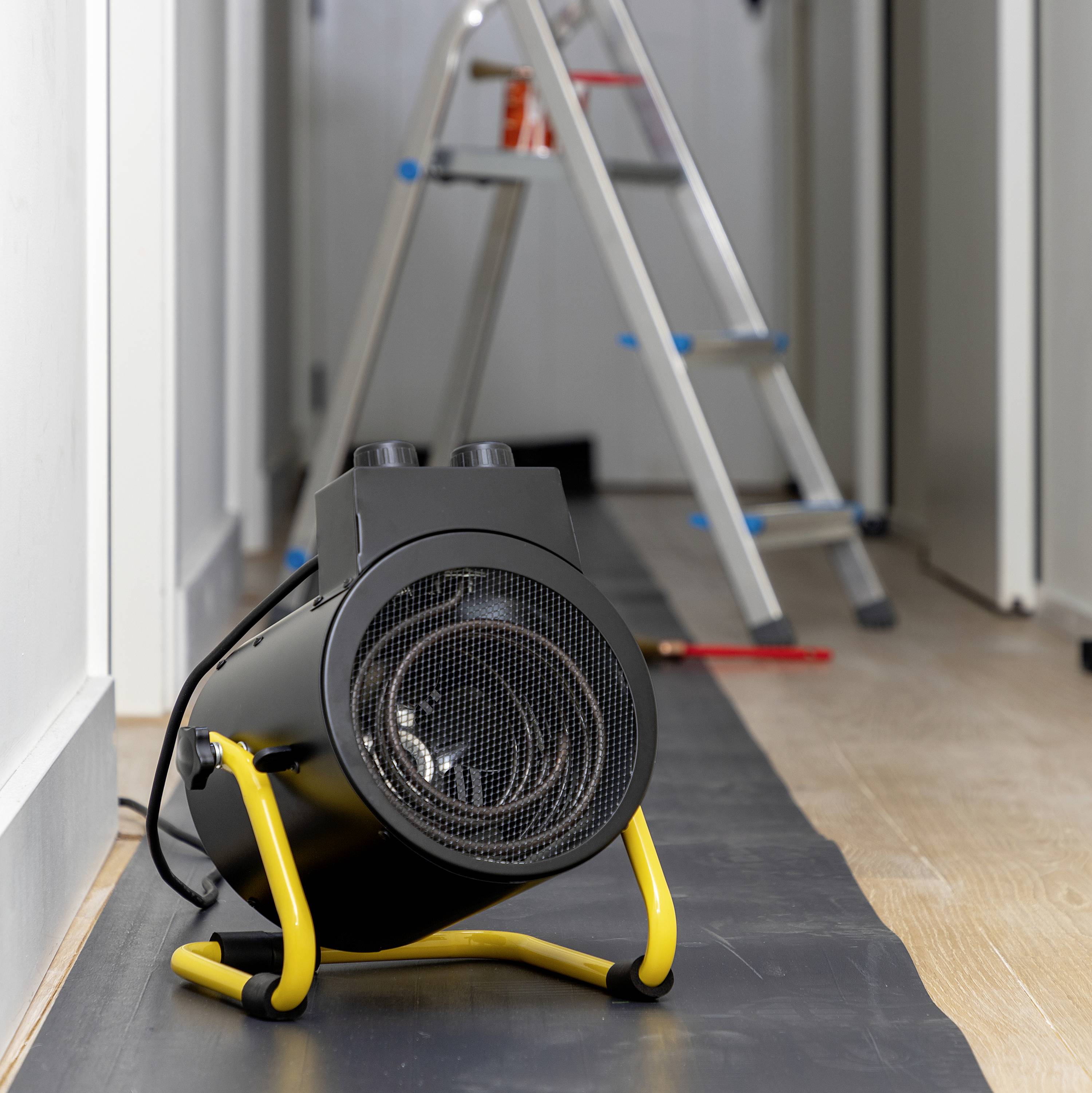 Radiateur électrique posé sur le sol d'un couloir, avec une échelle télescopique en arrière-plan. La scène suggère des travaux de rénovation en cours.