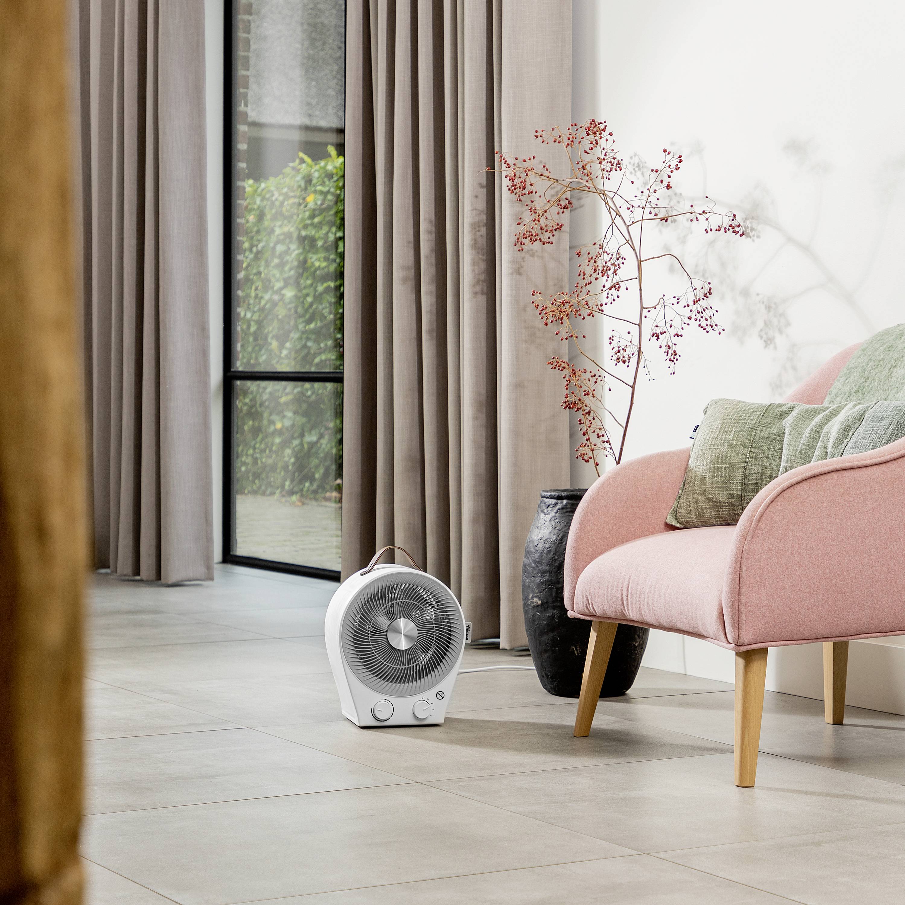 Un fauteuil rose est placé à côté d'une table et d'un vase contenant des branches. Un chauffage électrique d'appoint est posé sur le sol.