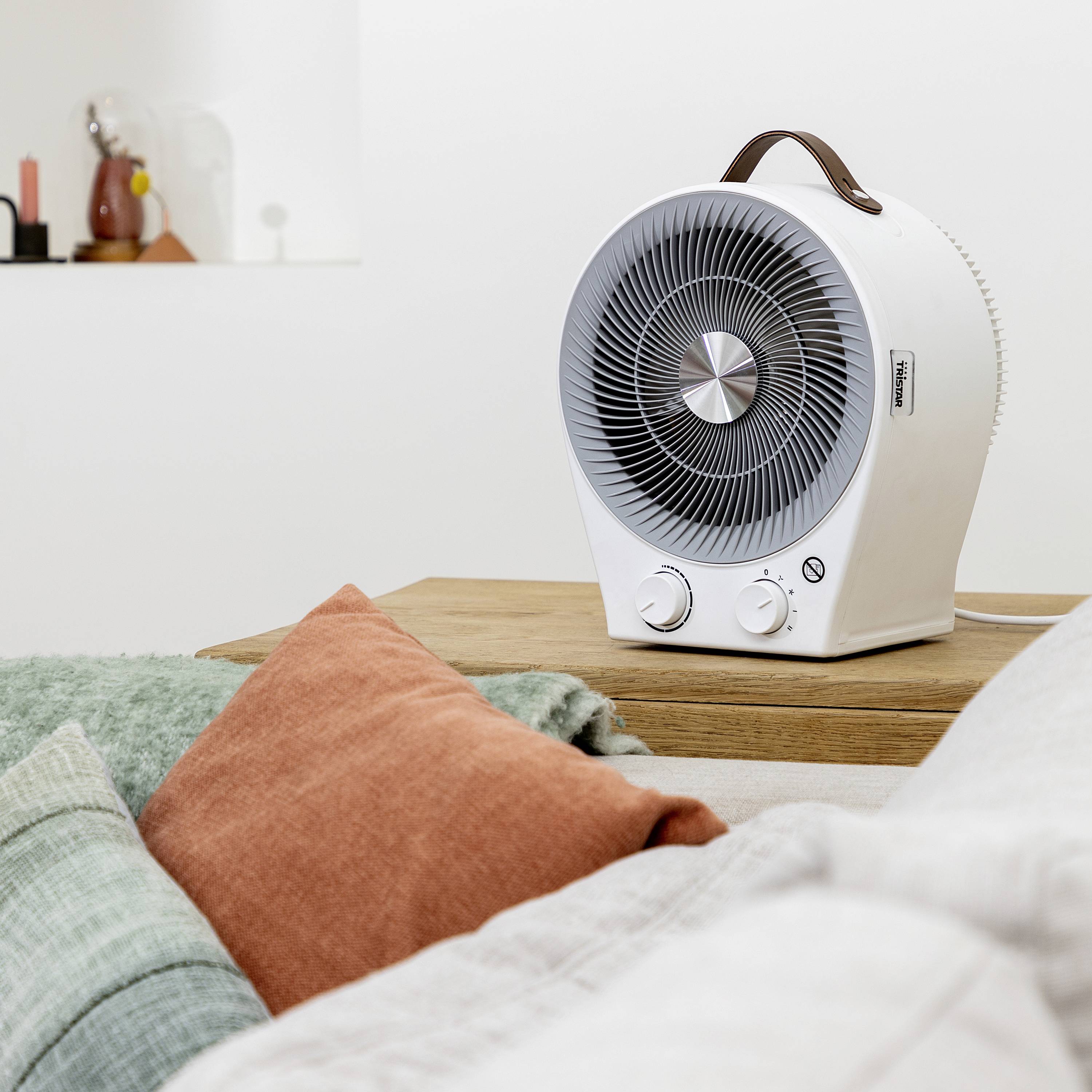 Un ventilateur chauffant blanc est posé sur une table en bois à côté de coussins de canapé aux tons terre. Des objets de décoration sont visibles en arrière-plan.