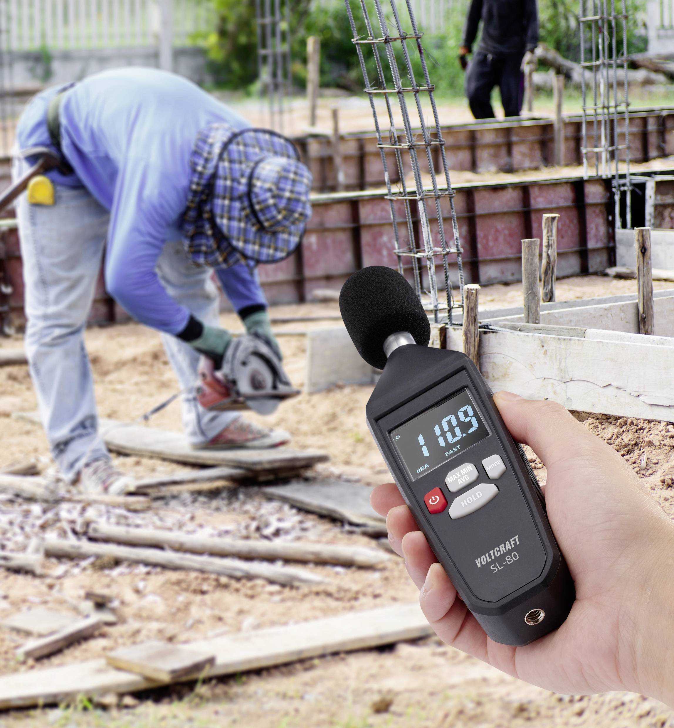Un appareil de mesure des décibels affiche '110,9', alors qu'en arrière-plan, une personne travaille avec une scie sur un chantier de construction.