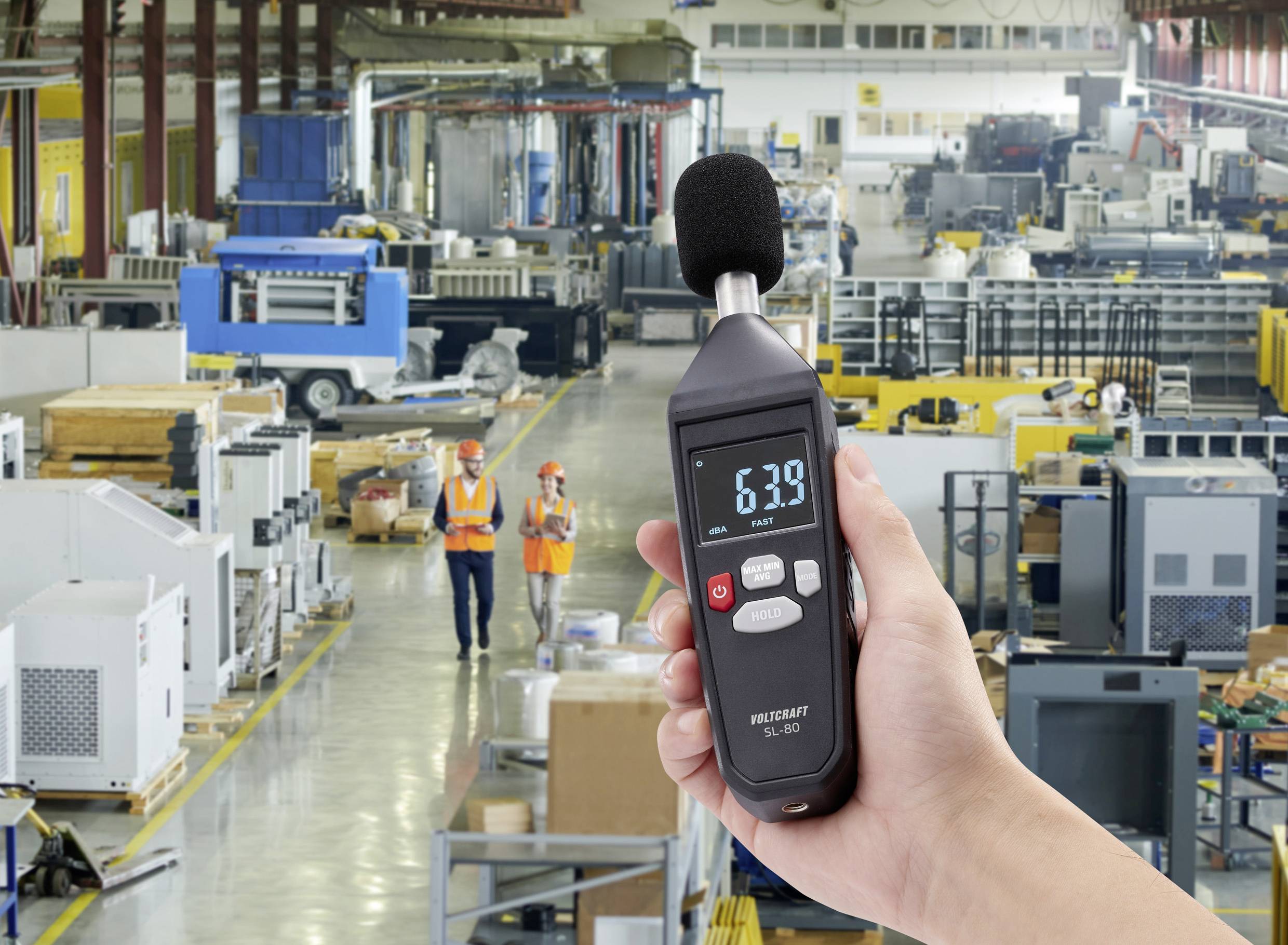 Une personne tient un sonomètre affichant 63,9 dB dans un grand environnement d'usine avec des machines et des employés en arrière-plan.