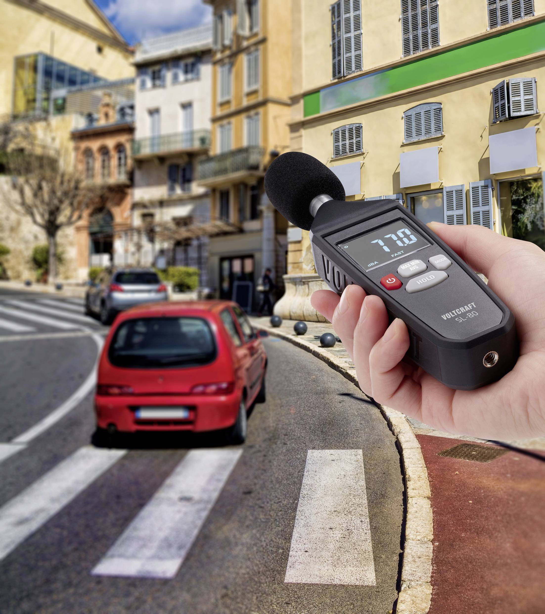 Une personne mesure le niveau sonore à l'aide d'un sonomètre près d'une rue animée avec une voiture rouge qui passe.