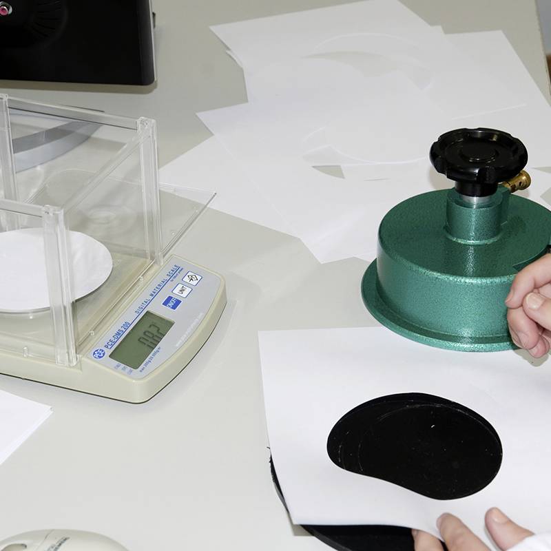 Une personne tient une feuille de papier blanche avec un cercle noir. À droite se trouve un appareil vert. Une balance électronique est située à gauche.
