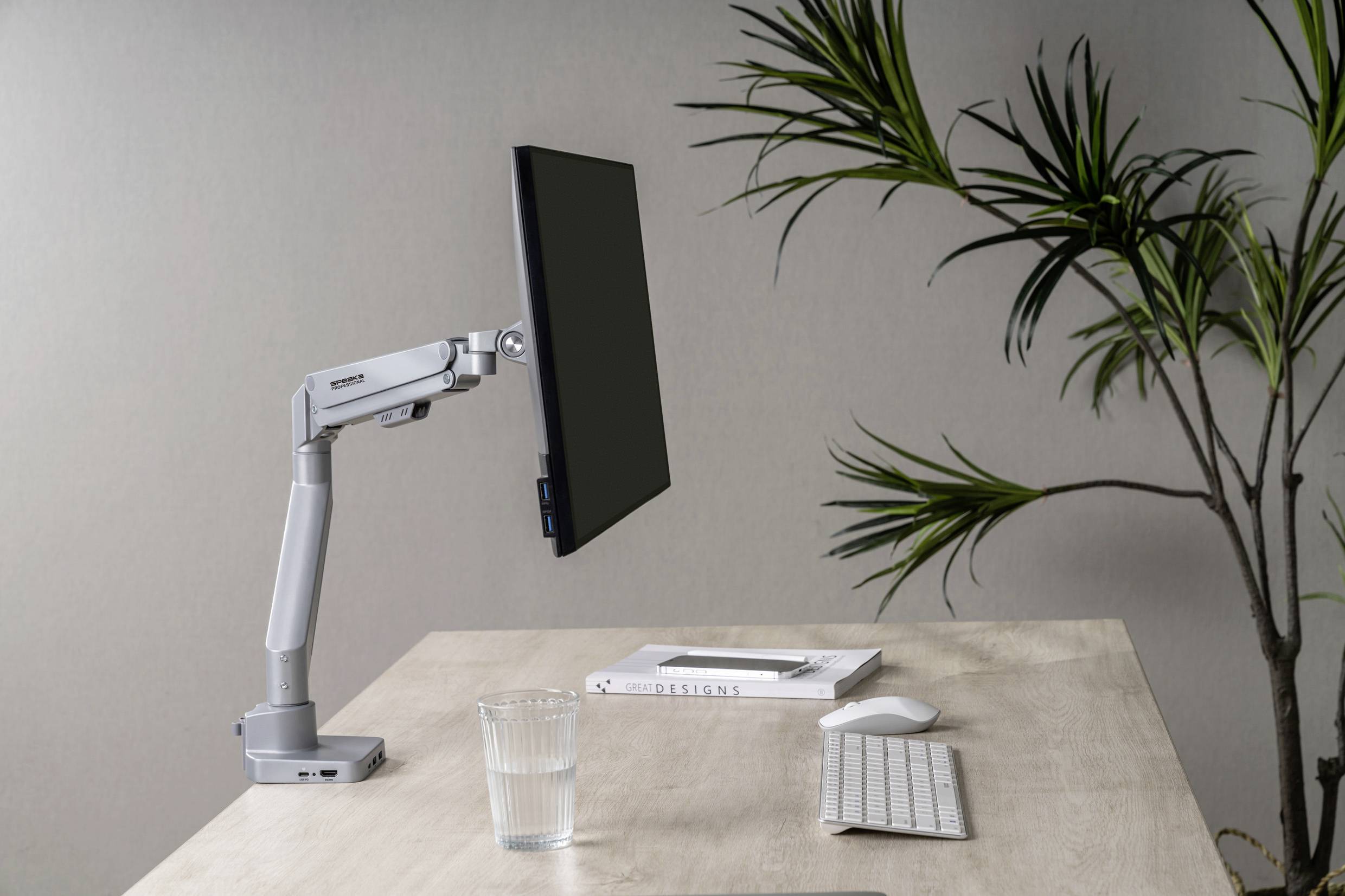 Un bureau moderne avec un bras de support d'écran assisté par ordinateur, un clavier, un verre d'eau et une plante en arrière-plan.
