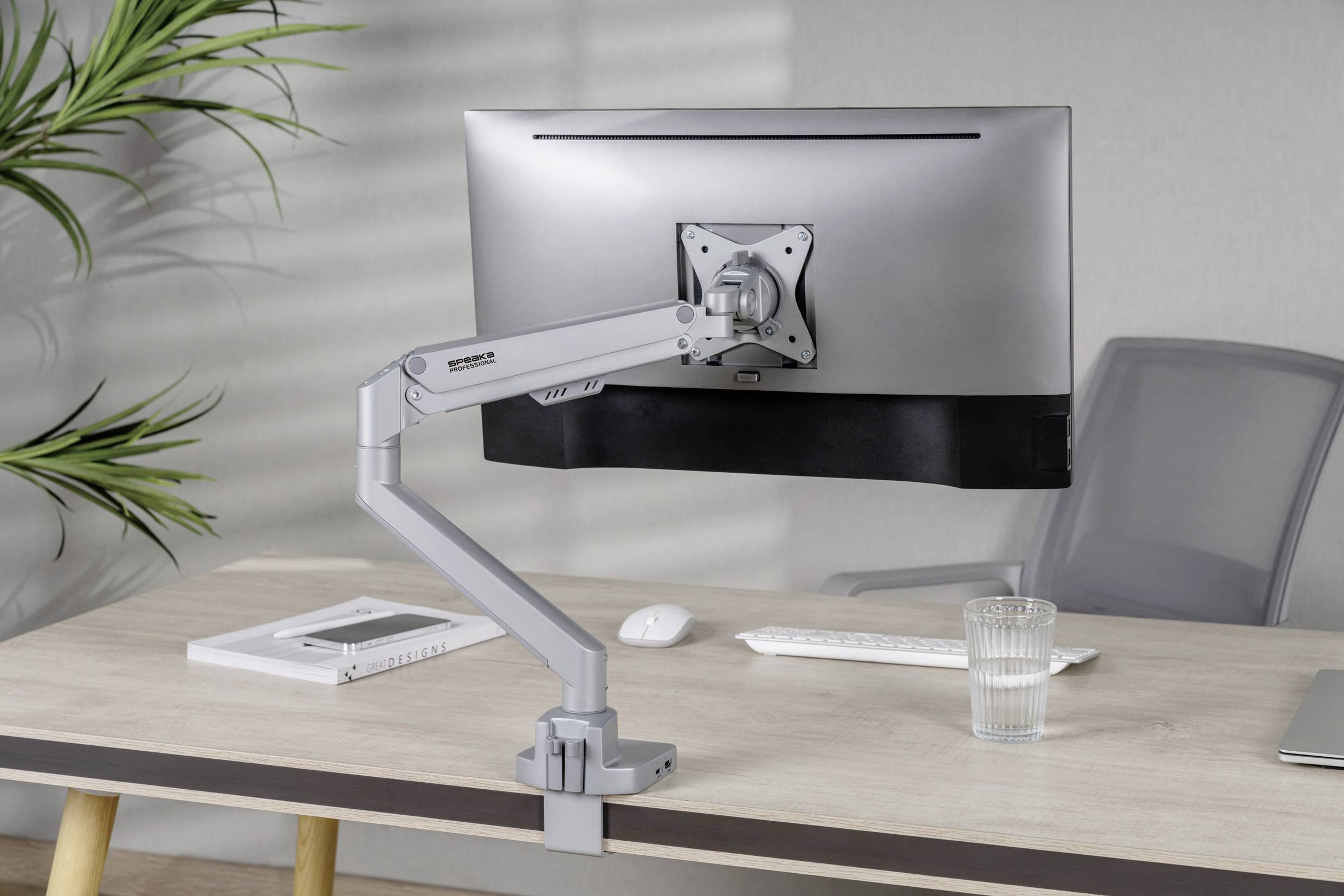 Un bureau avec un écran d'ordinateur sur un bras articulé, un clavier et un verre d'eau posés dessus ; en arrière-plan, un mur gris et une plante.