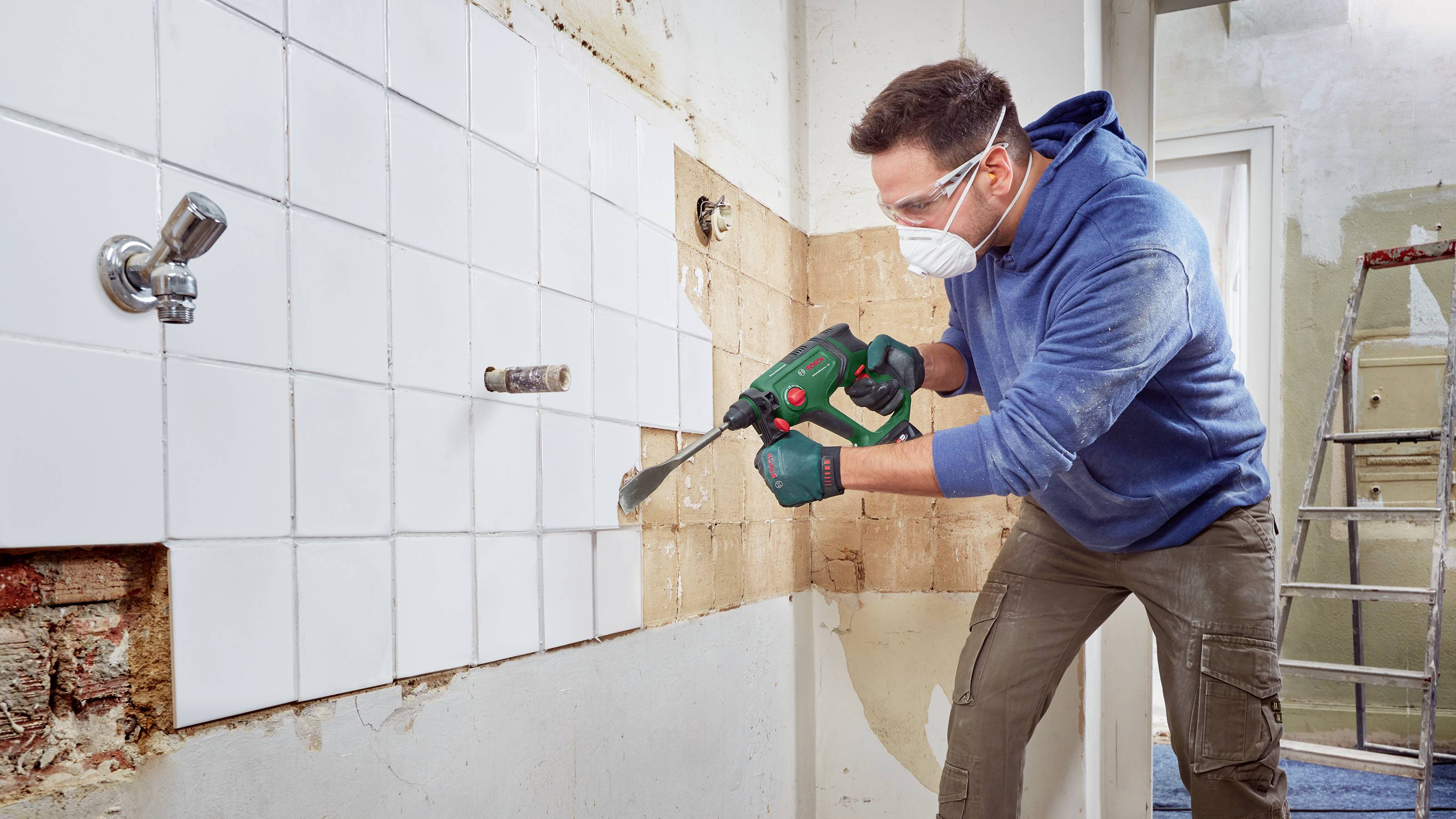 Un homme retire des carreaux blancs d'un mur à l'aide d'un outil électrique. Il porte des lunettes de protection et un masque. Travaux de rénovation.