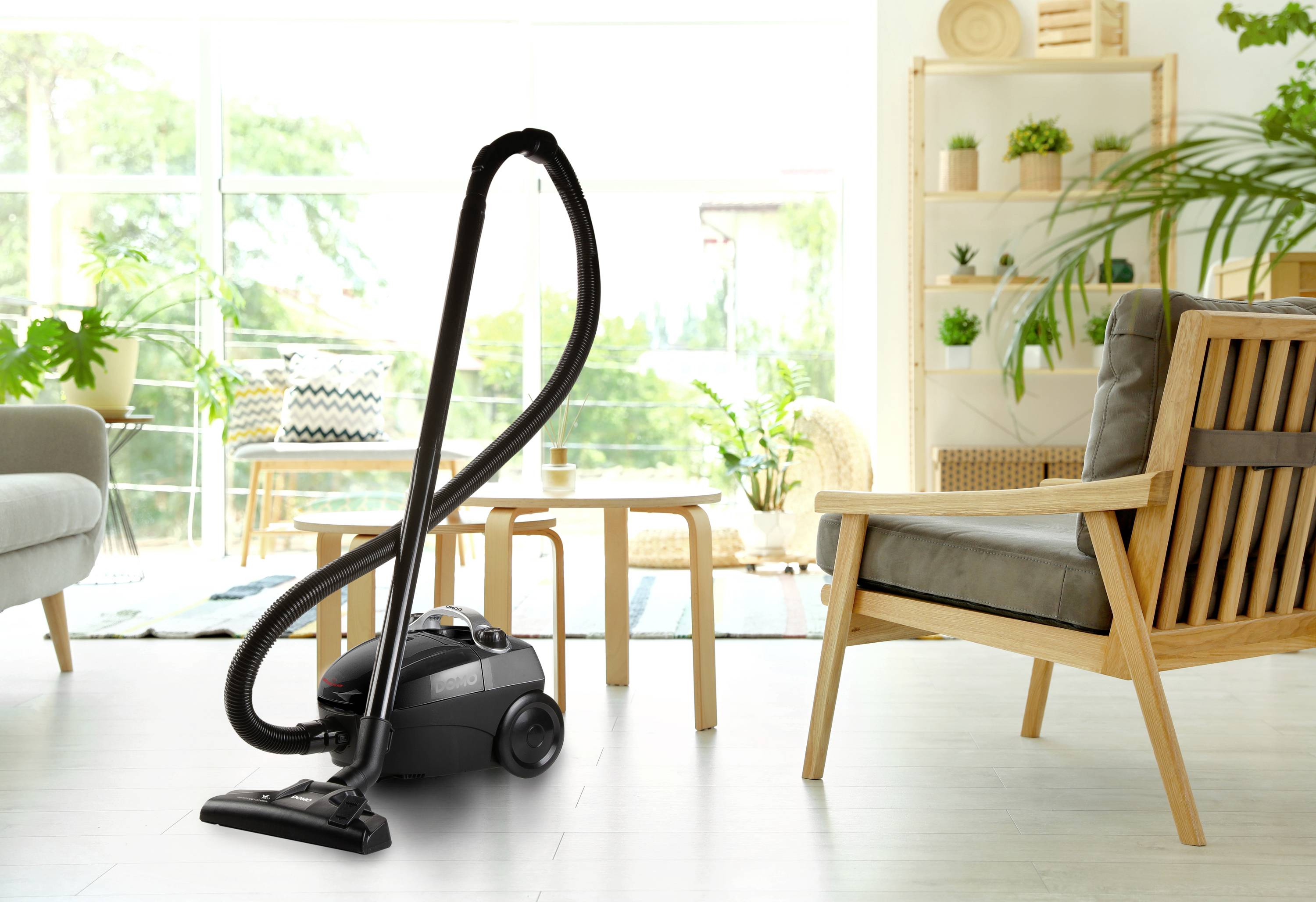 Un aspirateur est placé dans un salon moderne avec des meubles en bois et des plantes, sur un parquet clair, devant de grandes fenêtres.