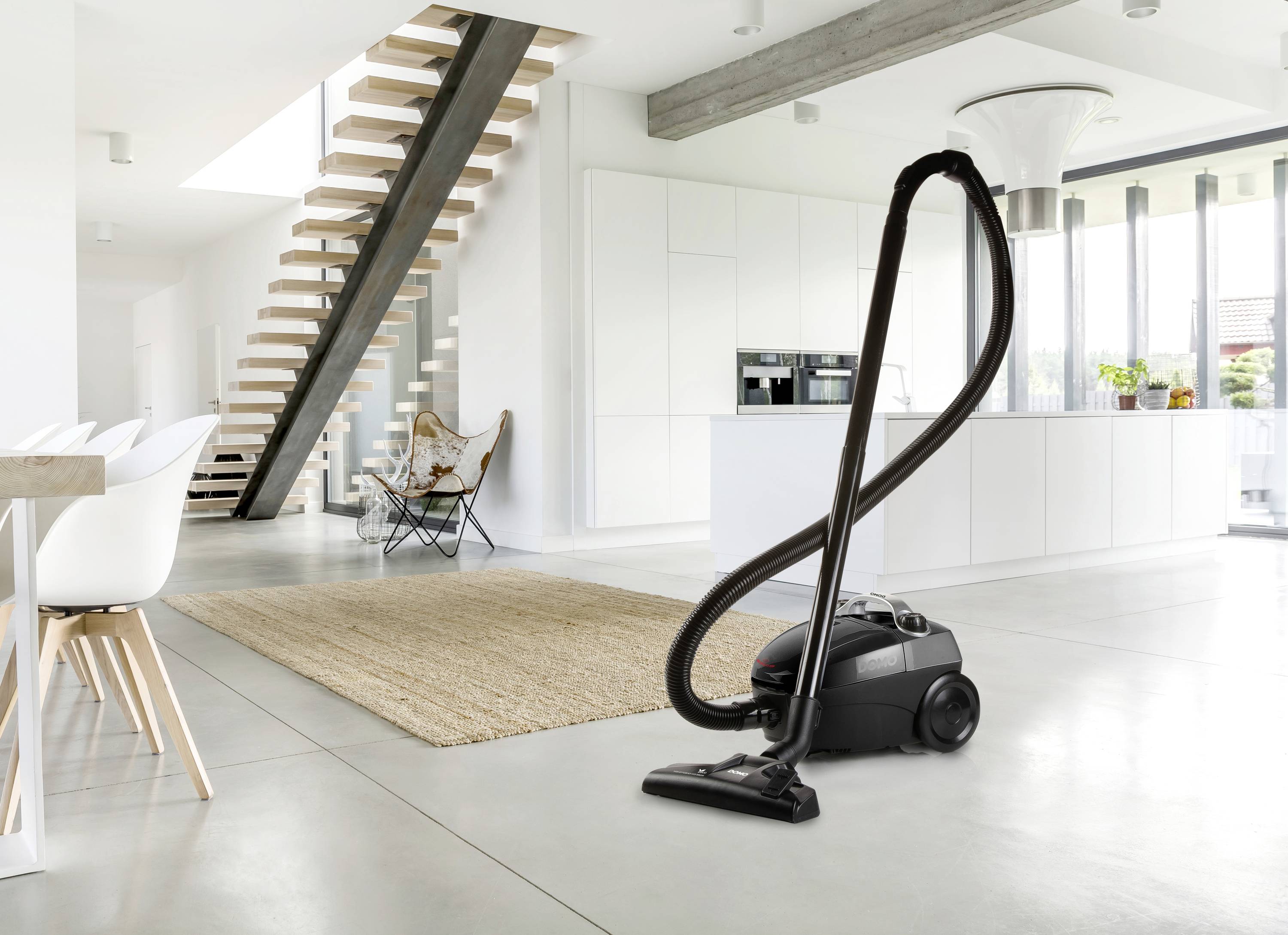 Un aspirateur noir est placé dans un salon moderne et lumineux avec des marches ouvertes et un grand tapis.