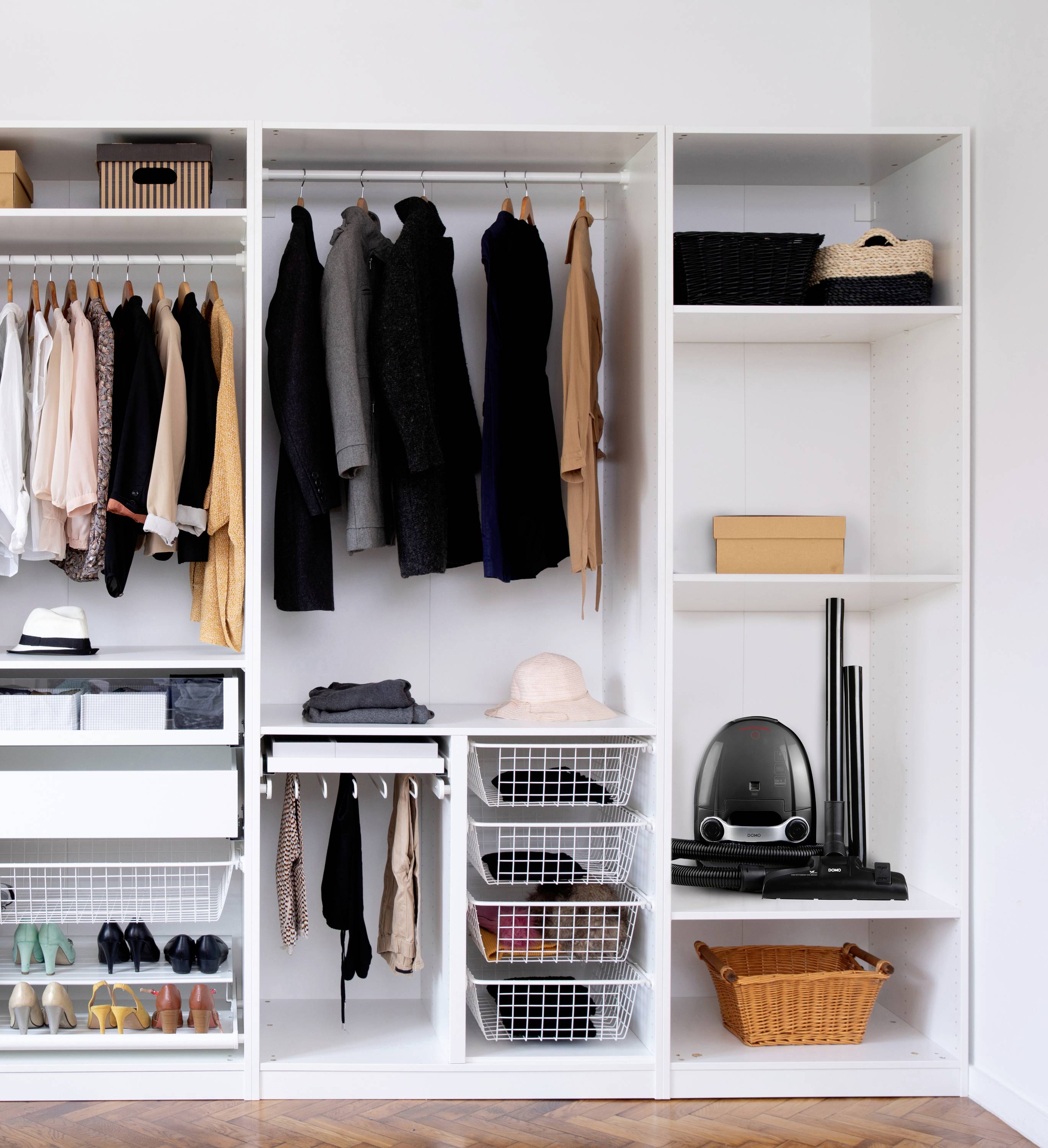 Armoire avec étagères suspendues, tiroirs et compartiments, remplie de vêtements, chaussures, chapeaux et un aspirateur à droite.
