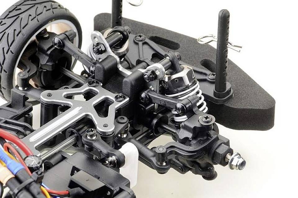 Gros plan sur l'essieu avant d'une voiture télécommandée, montrant l'amortisseur, la suspension et la roue avec des composants et un câblage visibles.