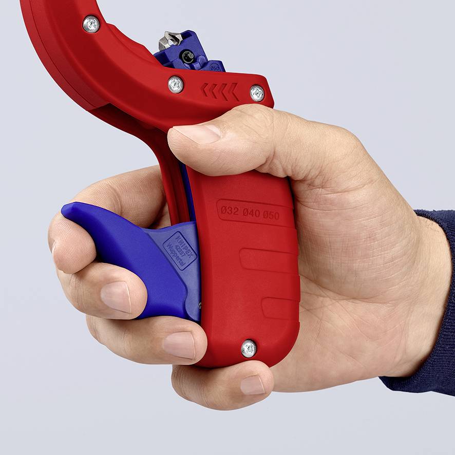 Une main tient un coupe-tube rouge avec une poignée bleue, prêt à couper des tuyaux. Le coupe-tube est équipé d'un mécanisme à cliquet intégré.