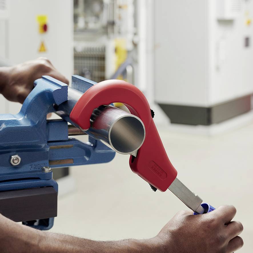 Une personne coupe un tuyau métallique avec une pince coupe-tube rouge, le tuyau étant fixé dans un étau bleu.