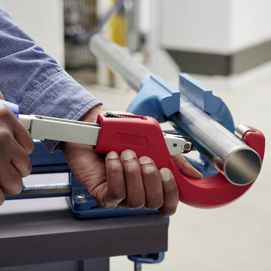 Une personne coupe un tuyau métallique avec un coupe-tube rouge. Le focus est mis sur la main et l'outil de coupe.