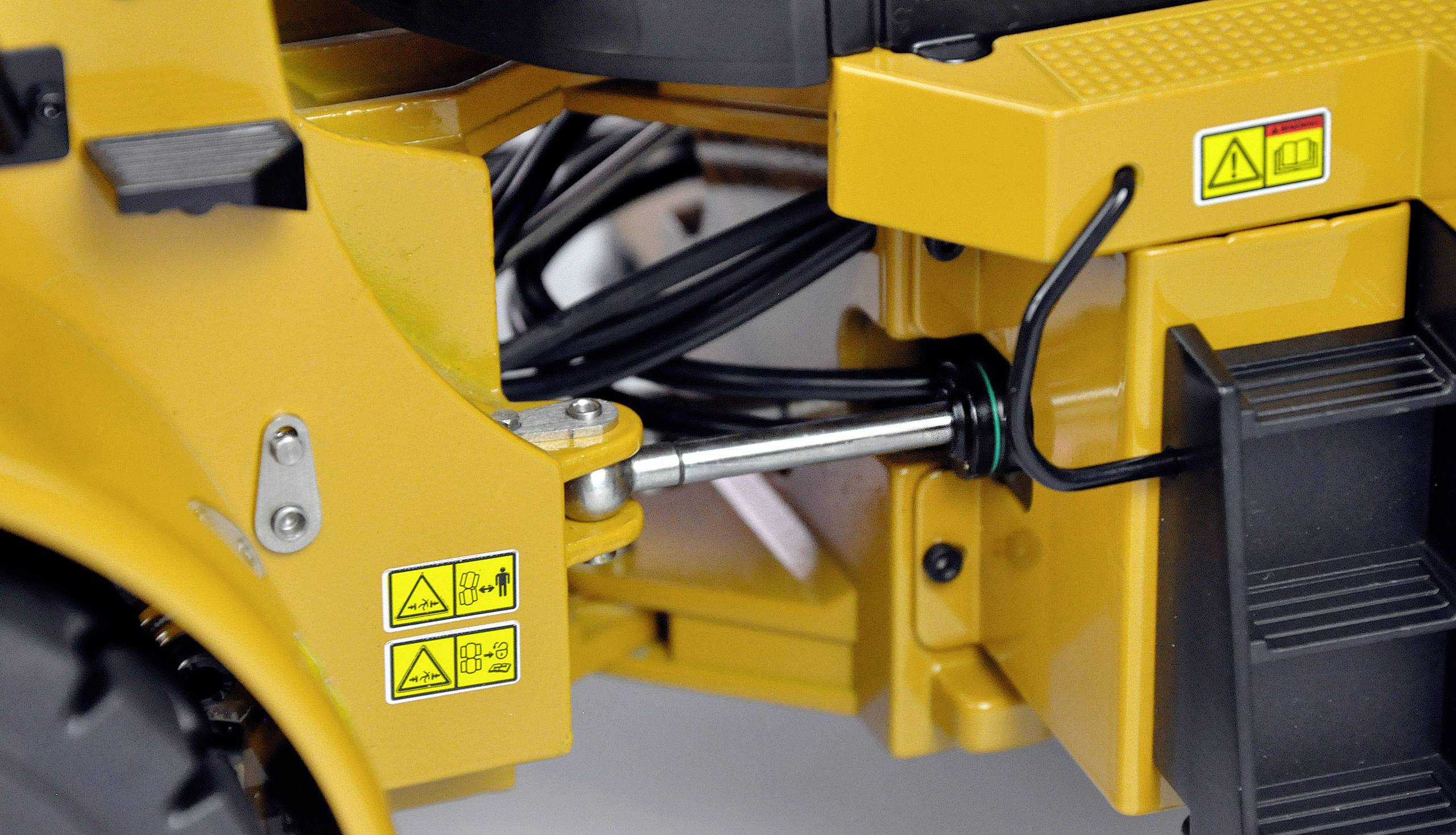 Gros plan d'un bras de machine industrielle jaune avec des conduites hydrauliques visibles et des symboles d'avertissement.