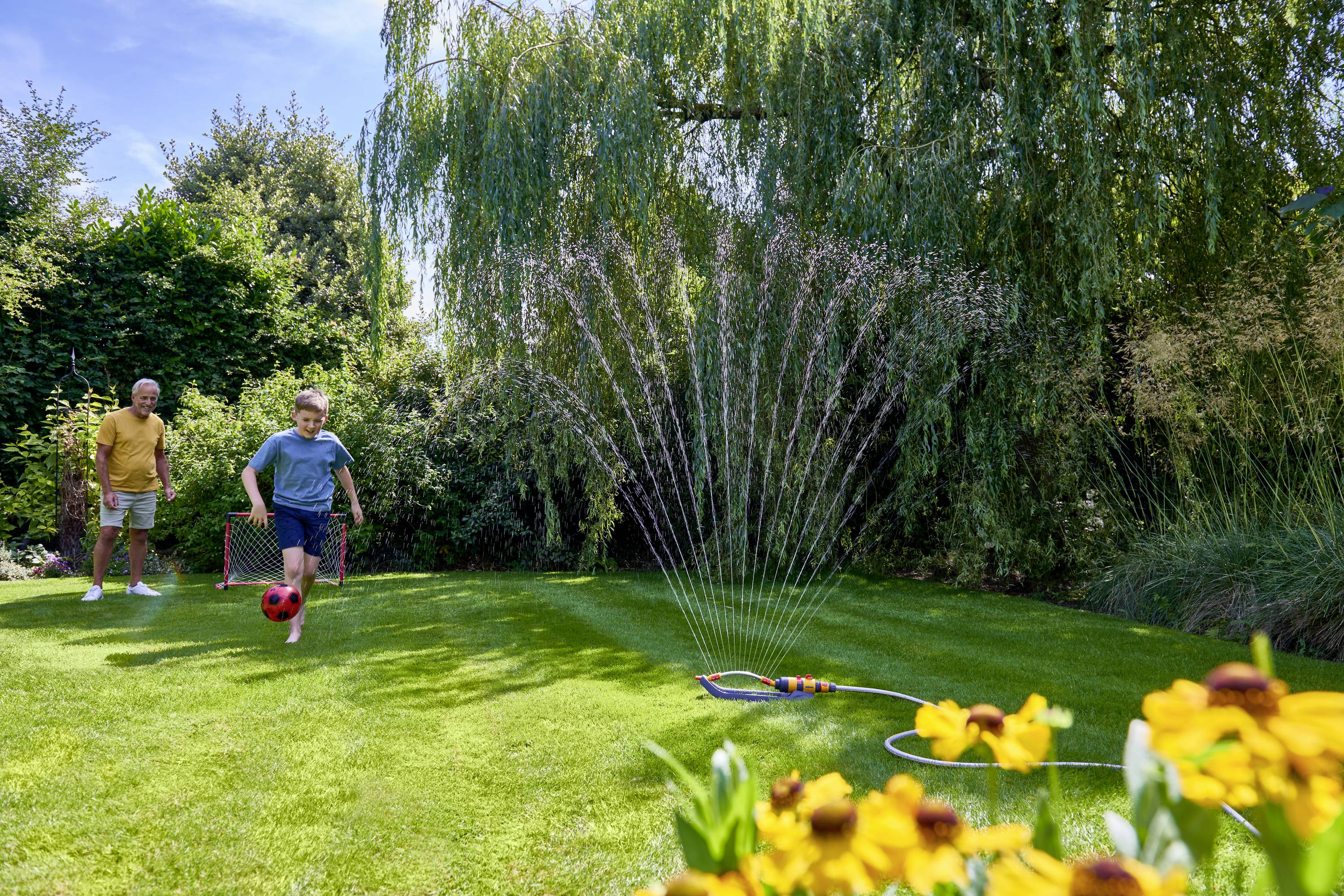 Un enfant et un adulte jouent au football dans le jardin. Un arroseur automatique arrose la pelouse, entouré d'arbres et de fleurs.
