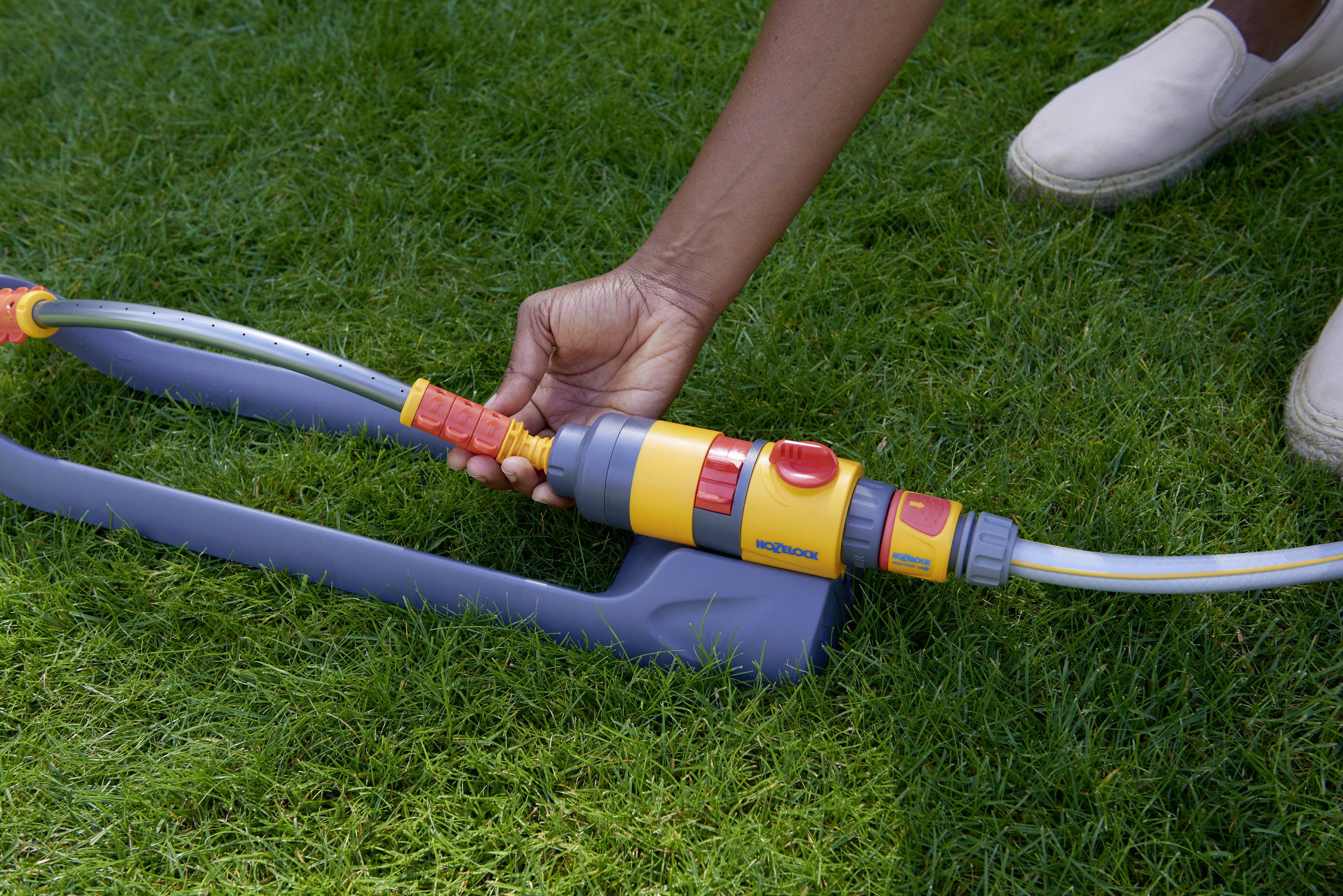 Une main raccorde un tuyau d'arrosage à un arroseur automatique sur de l'herbe verte.