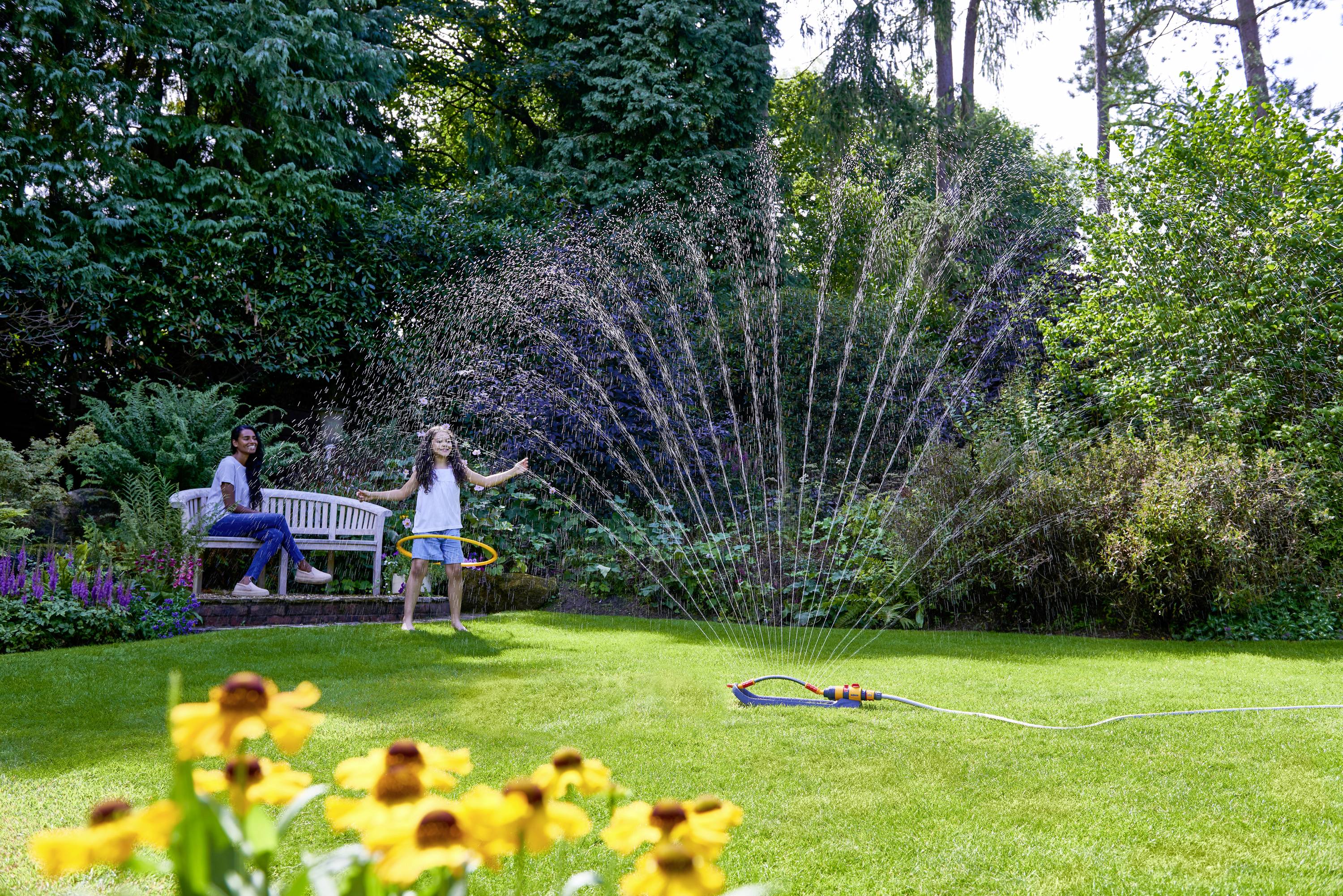 Deux enfants joyeux jouent avec un arroseur dans le jardin sous le soleil. Une personne est assise sur un banc en arrière-plan.