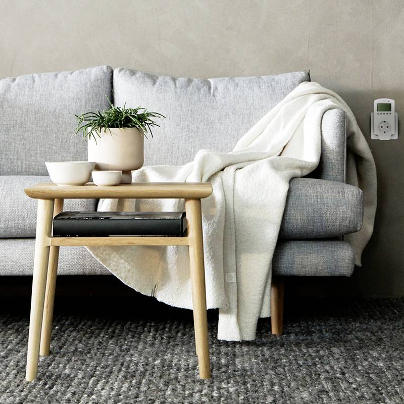 Un salon moderne avec un canapé gris, un jeté beige, une table en bois avec une plante, des tasses et un sol moquetté.