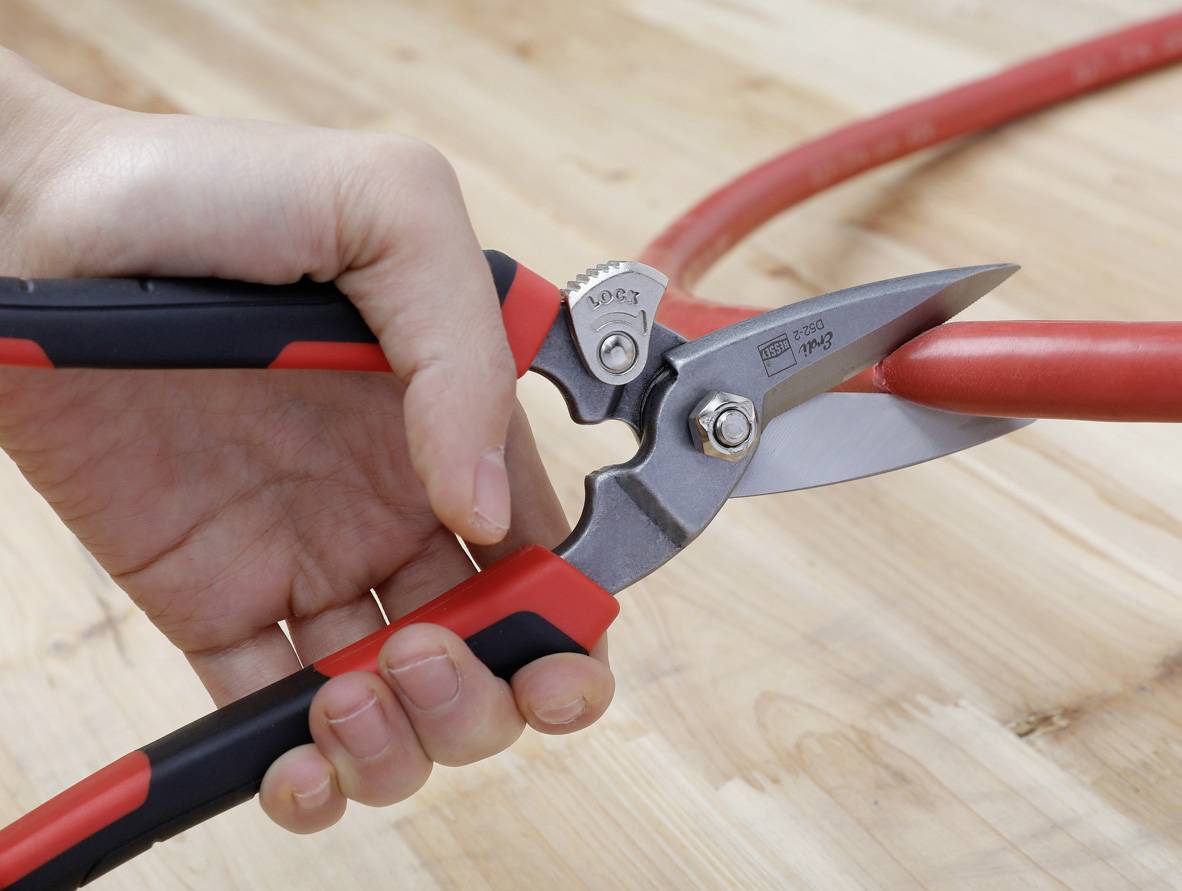 Une main tient un sécateur qui coupe un câble rouge. La lame est tranchante et se referme fermement autour du câble.