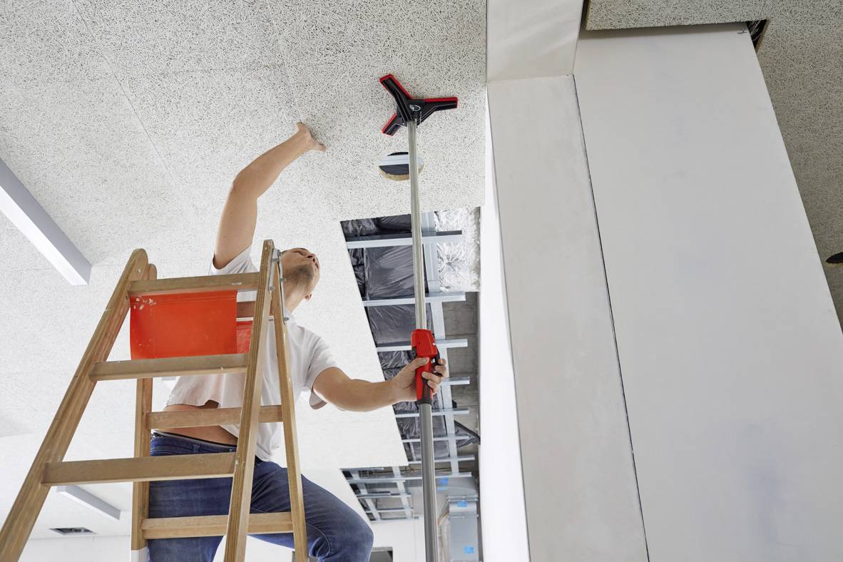 Une personne sur une échelle répare un plafond suspendu à l'aide d'un long outil dans un immeuble de bureaux.