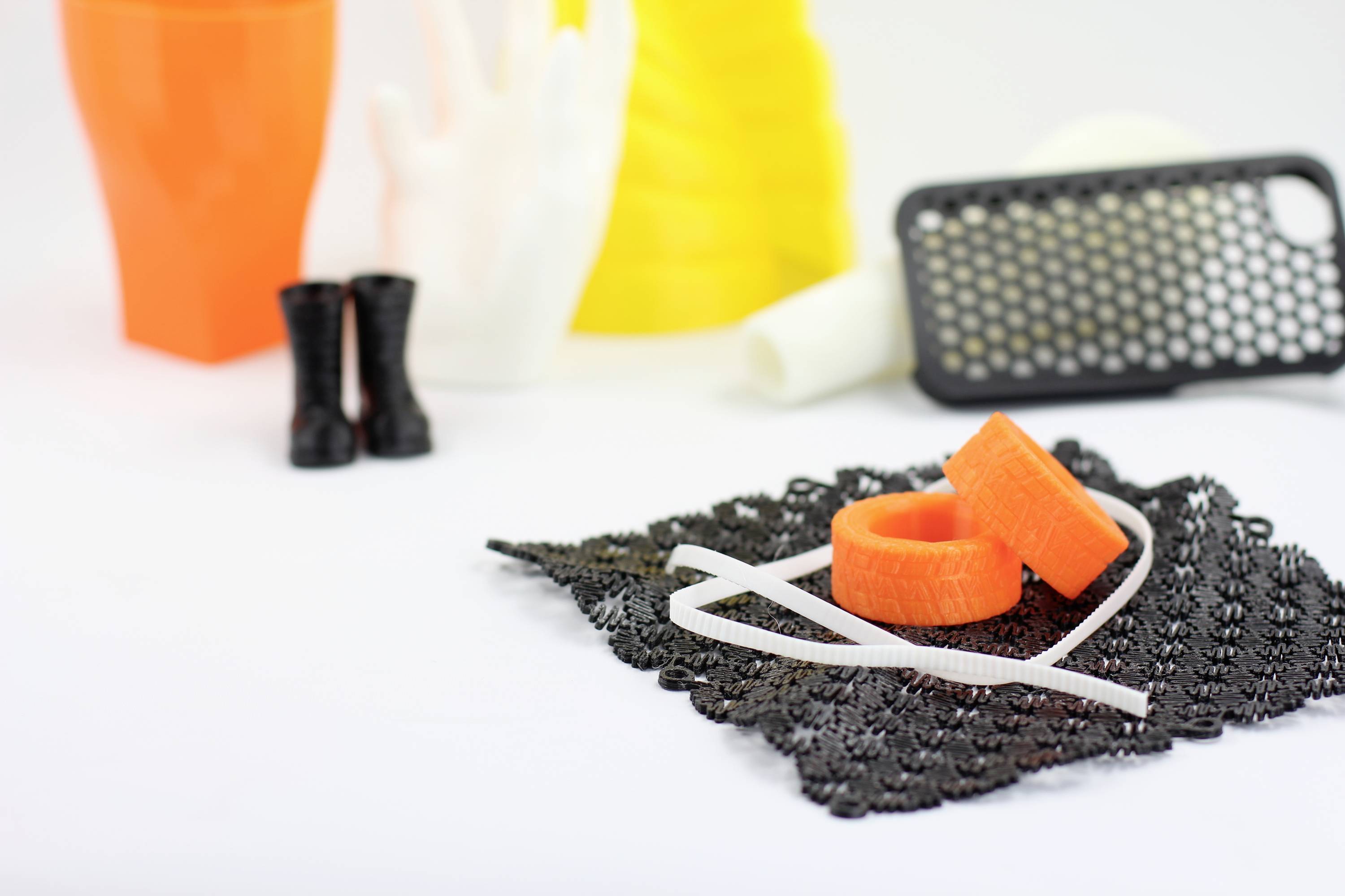 Objets imprimés en 3D : Deux anneaux oranges et des bandes blanches sur un fond noir mat. En arrière-plan : chaussures, vases, étui de téléphone.
