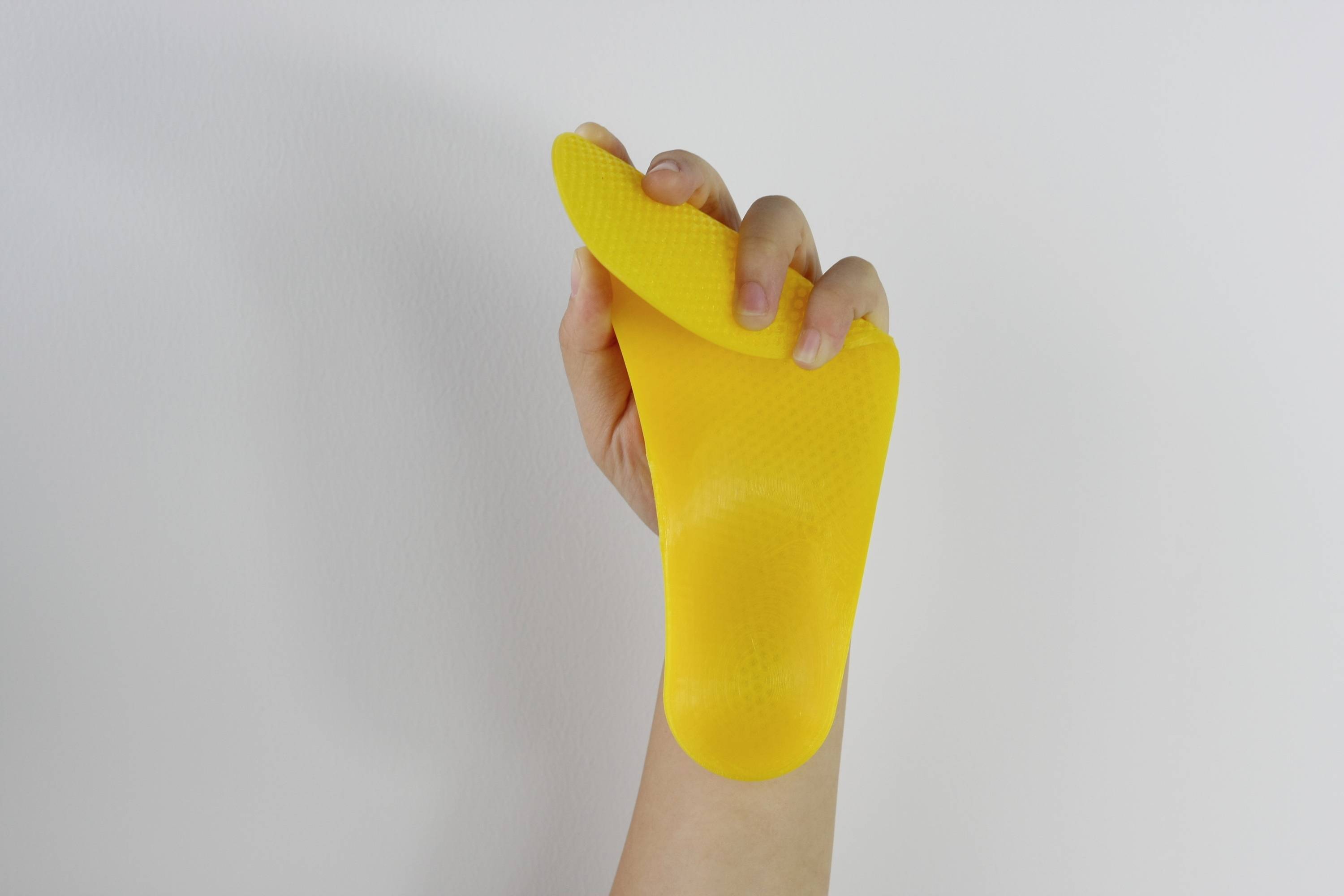 Une main tient un ustensile de cuisine jaune et flexible, probablement un gant de four ou un protège-chaleur, contre un fond neutre.