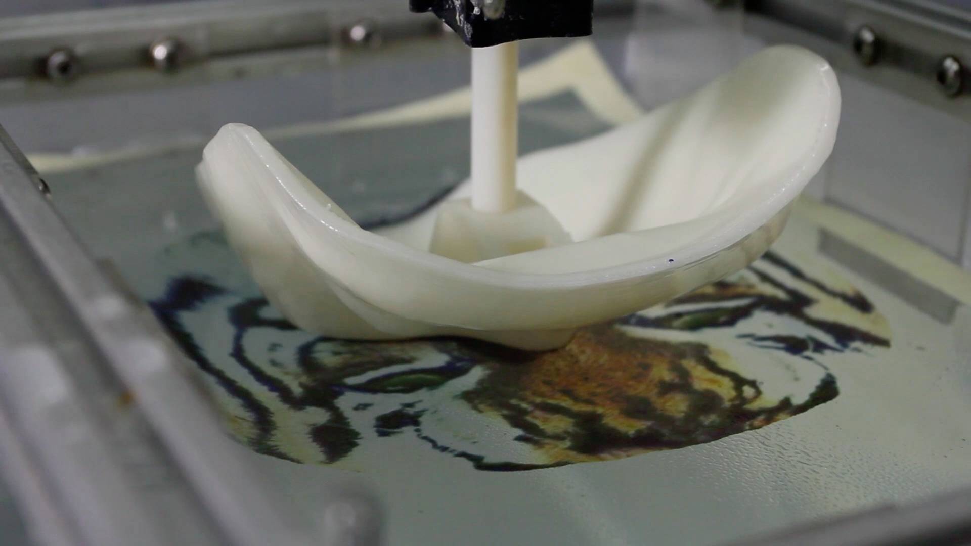 Un imprimeur 3D forme un objet incurvé. En arrière-plan, on peut voir l'image d'un tigre sur un écran.