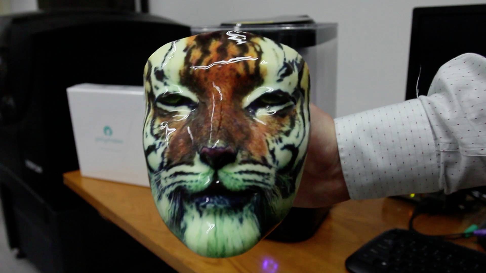 Une main tient un masque représentant un visage de tigre. Des appareils de bureau sont visibles en arrière-plan.