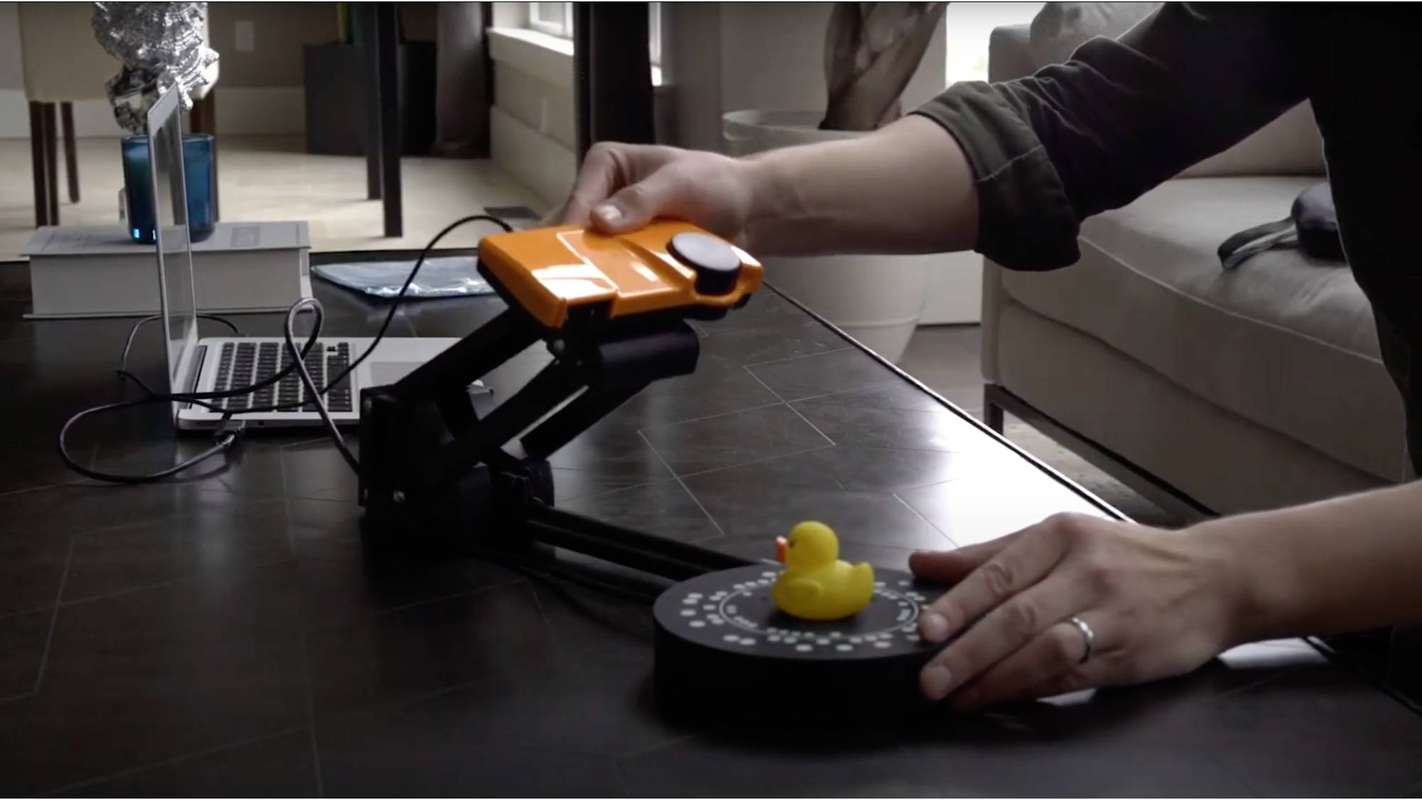 Une personne numérise un canard en caoutchouc jaune sur un plateau tournant rond à l'aide d'un scanner 3D orange, posé sur une table.