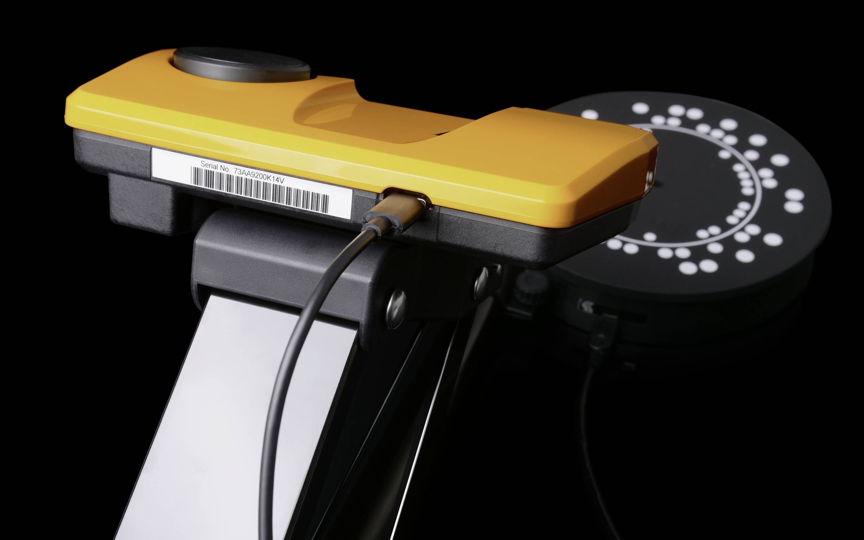 Un appareil jaune et noir avec une connexion filaire sur un trépied, devant un fond noir à motif pointillé.