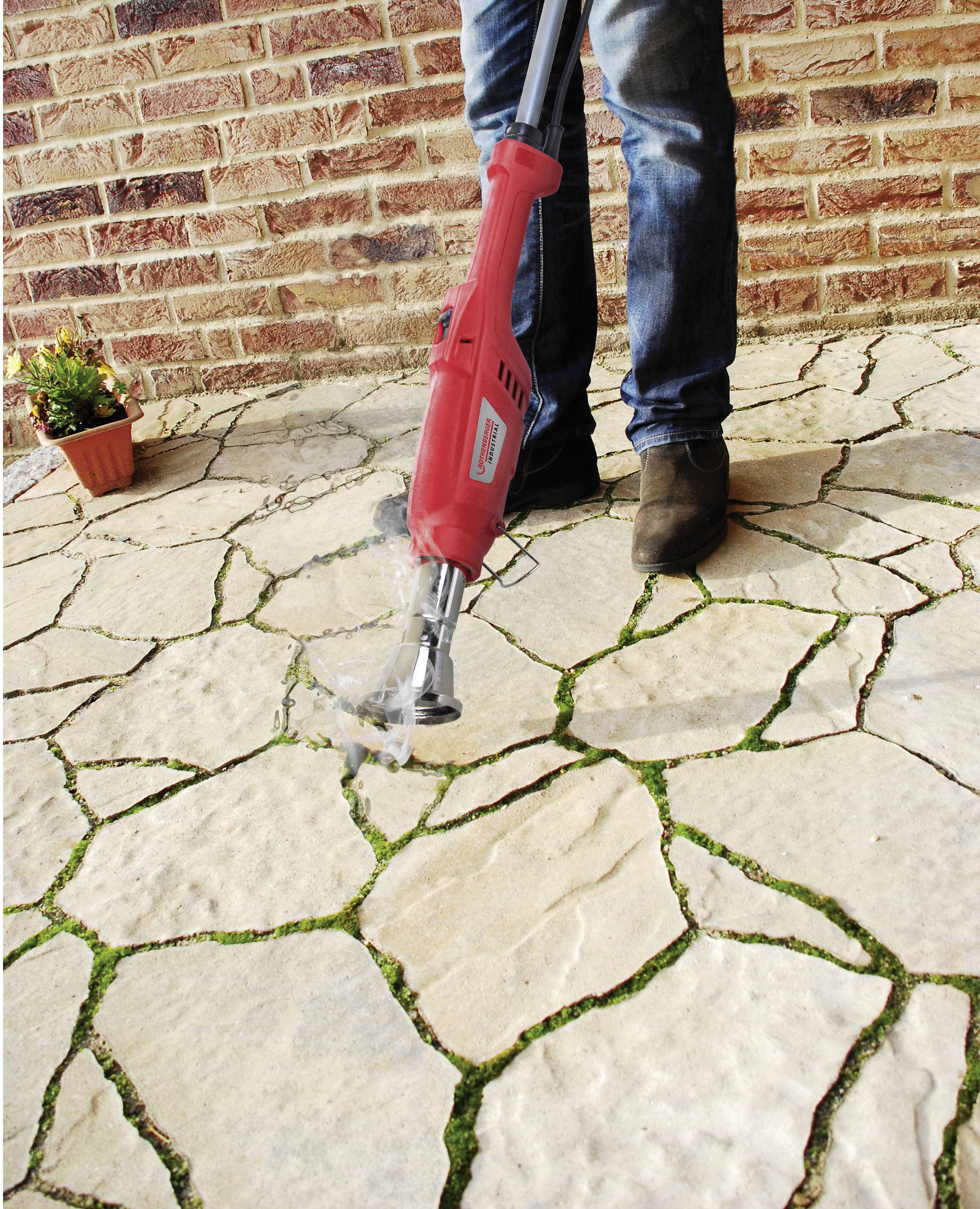 Une personne retire des mauvaises herbes avec un dispositif rouge entre les joints d'un chemin pavé devant un mur de briques.
