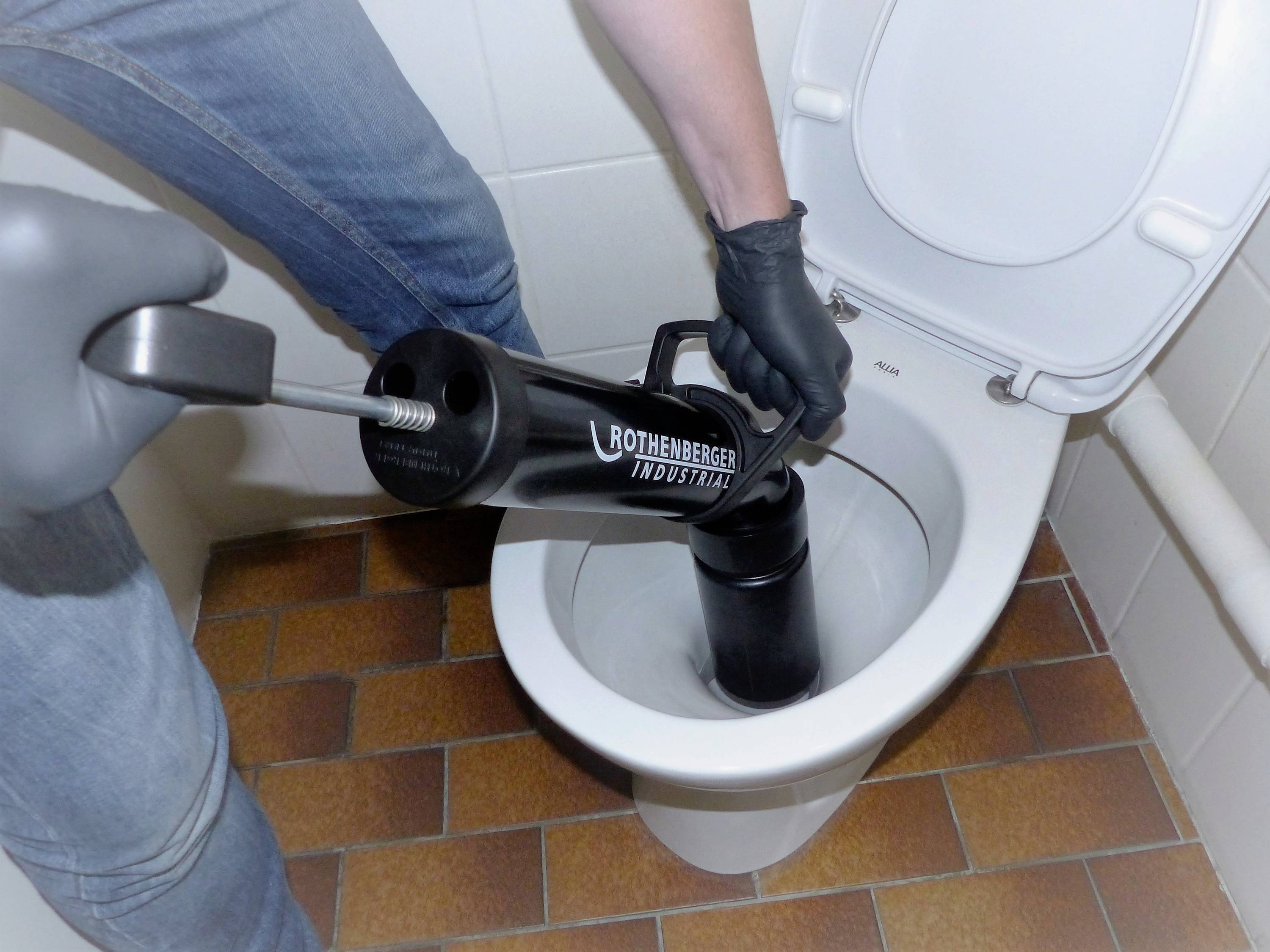 Une personne nettoie des toilettes avec un déboucheur manuel dans une salle de bain.