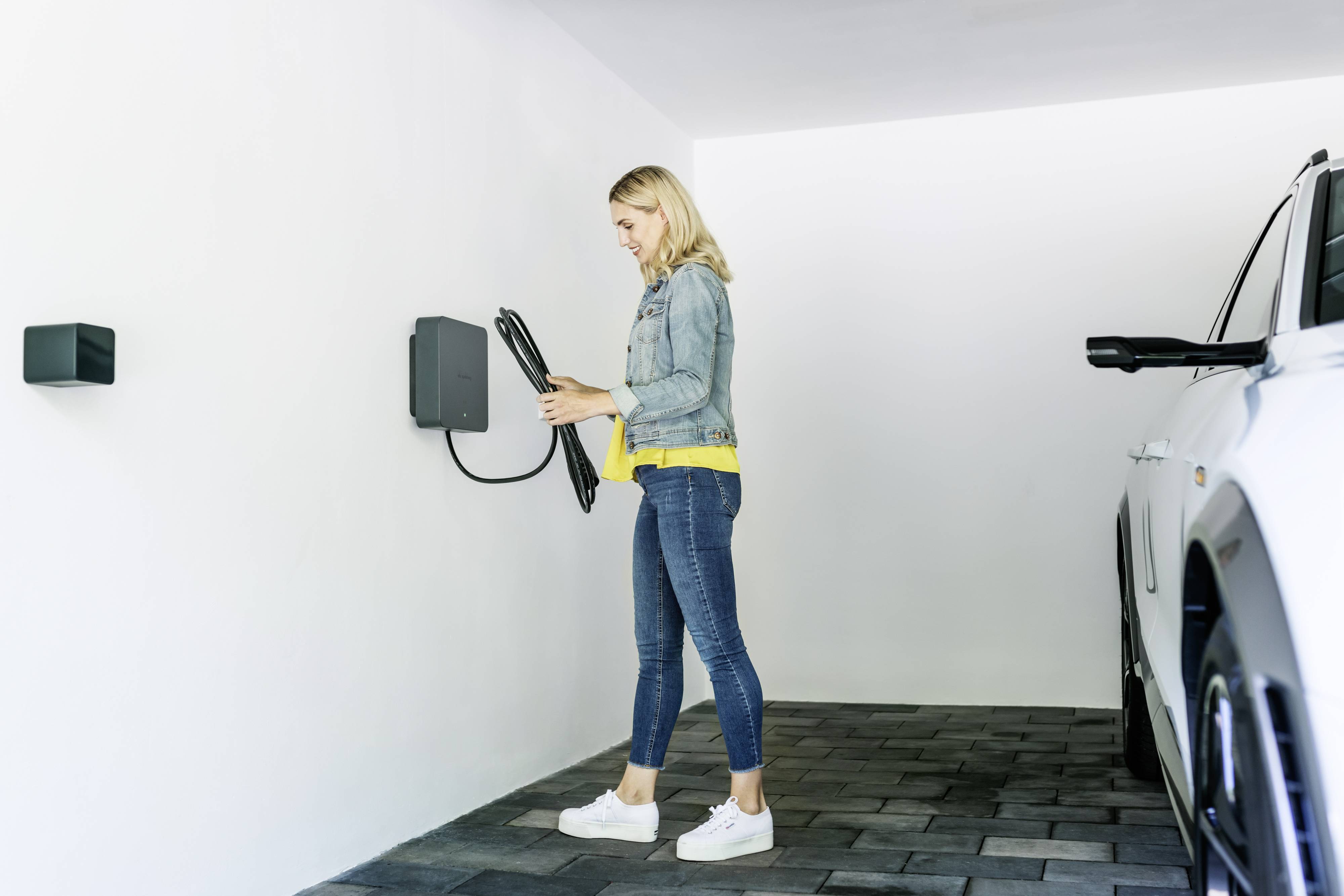 Une femme portant un jean et un chemisier jaune se tient dans un garage et tient un câble de charge à côté d'une voiture électrique.