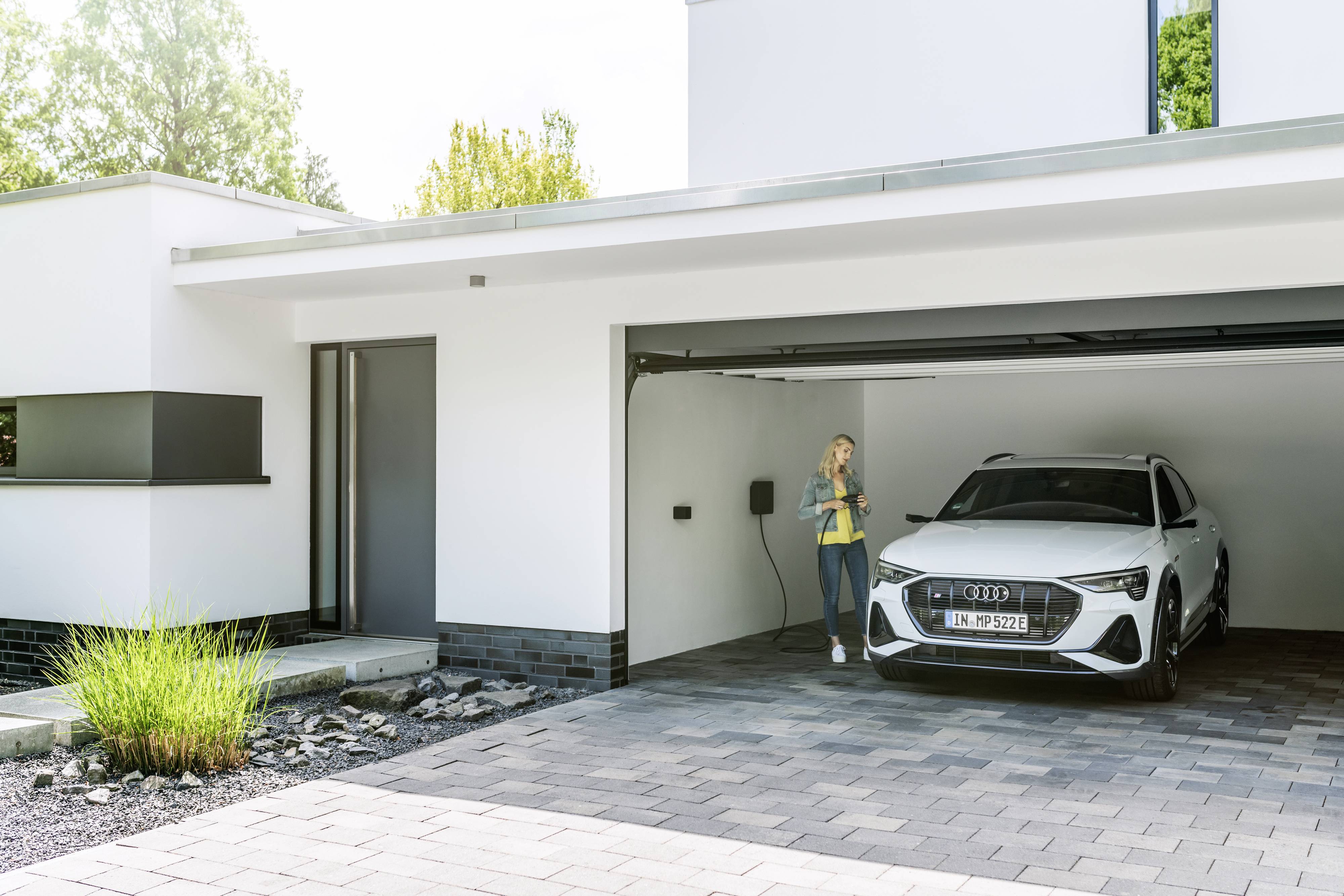 Une femme charge une voiture électrique dans un garage moderne. La voiture est blanche et se trouve sous une porte de garage ouverte.