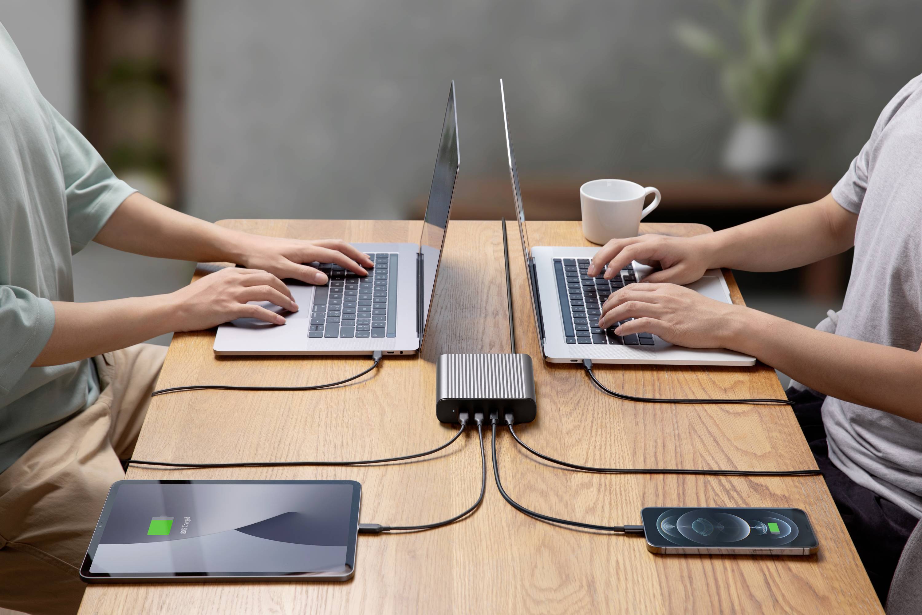 Deux personnes travaillent sur des ordinateurs portables à une table, branchés à un chargeur. Une tablette et un smartphone sont chargés simultanément.
