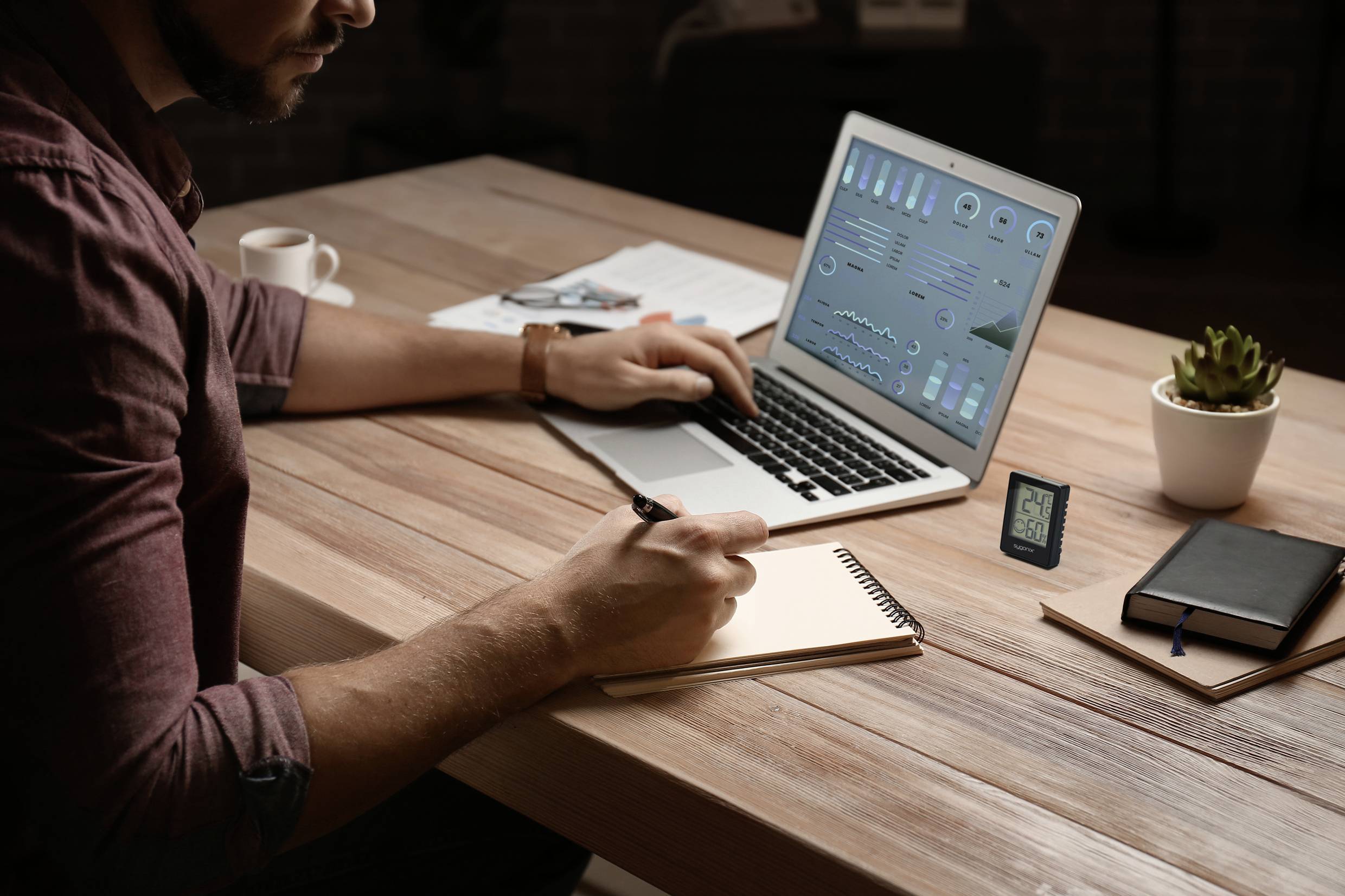 Un homme est assis à une table en bois, utilise un ordinateur portable affichant des données à l'écran et prend des notes dans un carnet.