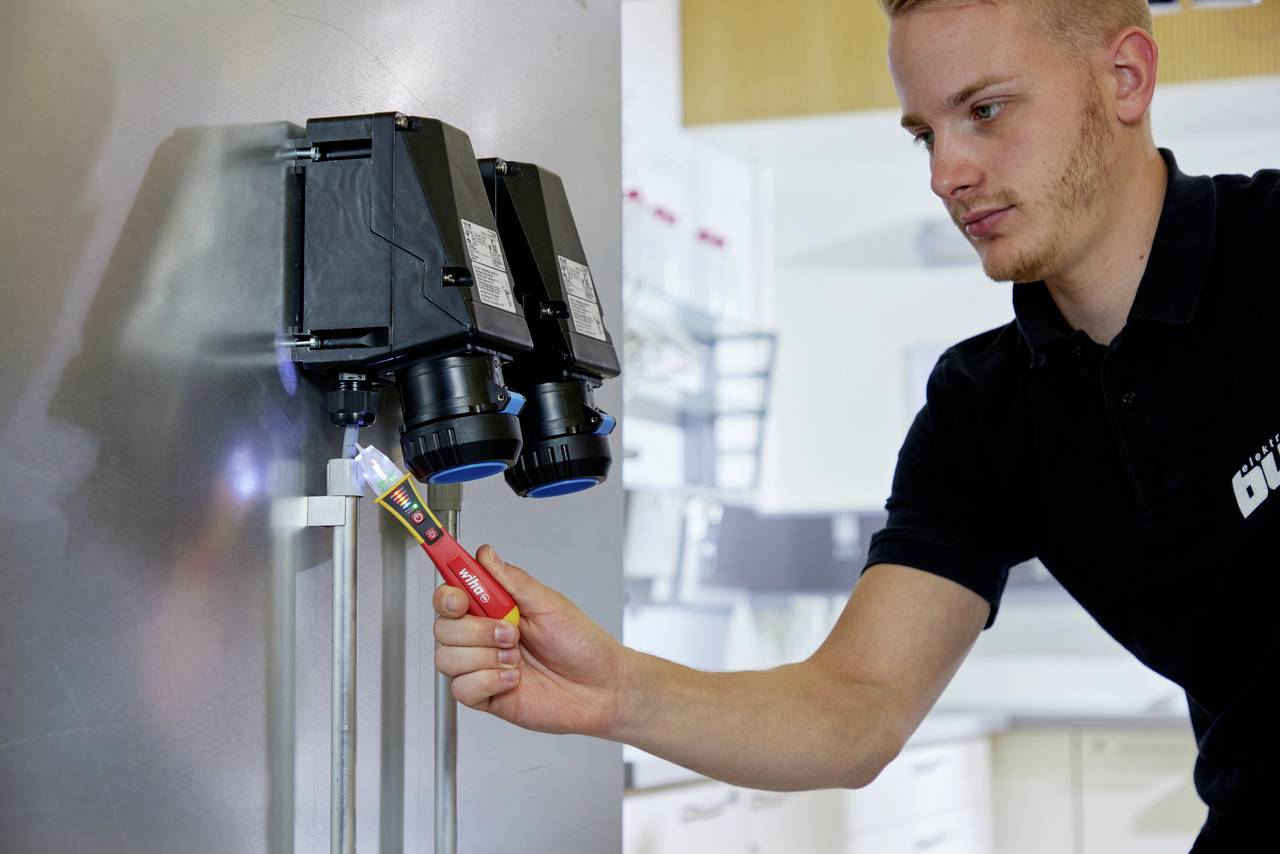 Un technicien en polo noir vérifie l'électricité d'une machine à l'aide d'un testeur de tension dans un atelier industriel.