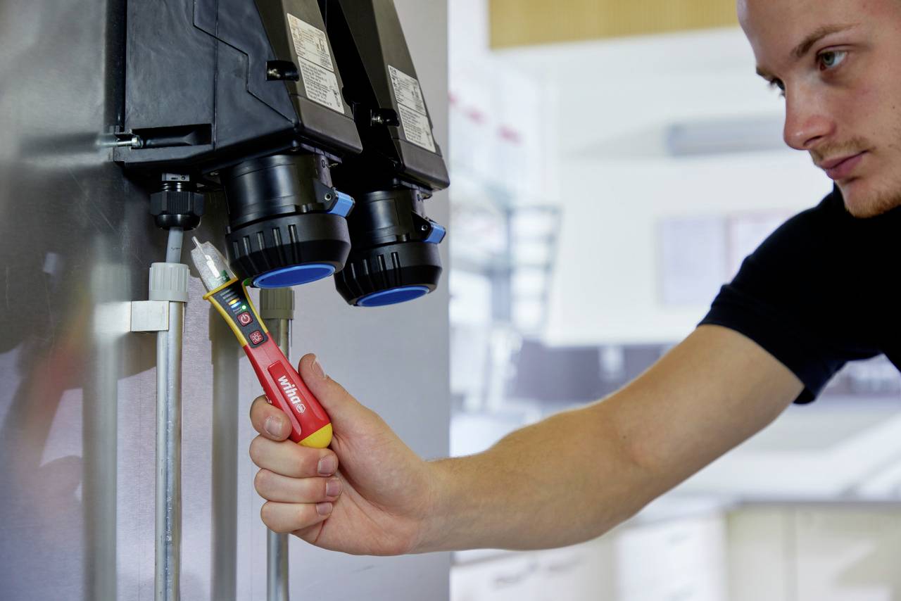 Un homme utilise un testeur de tension sur une installation électrique. Son expression est concentrée.