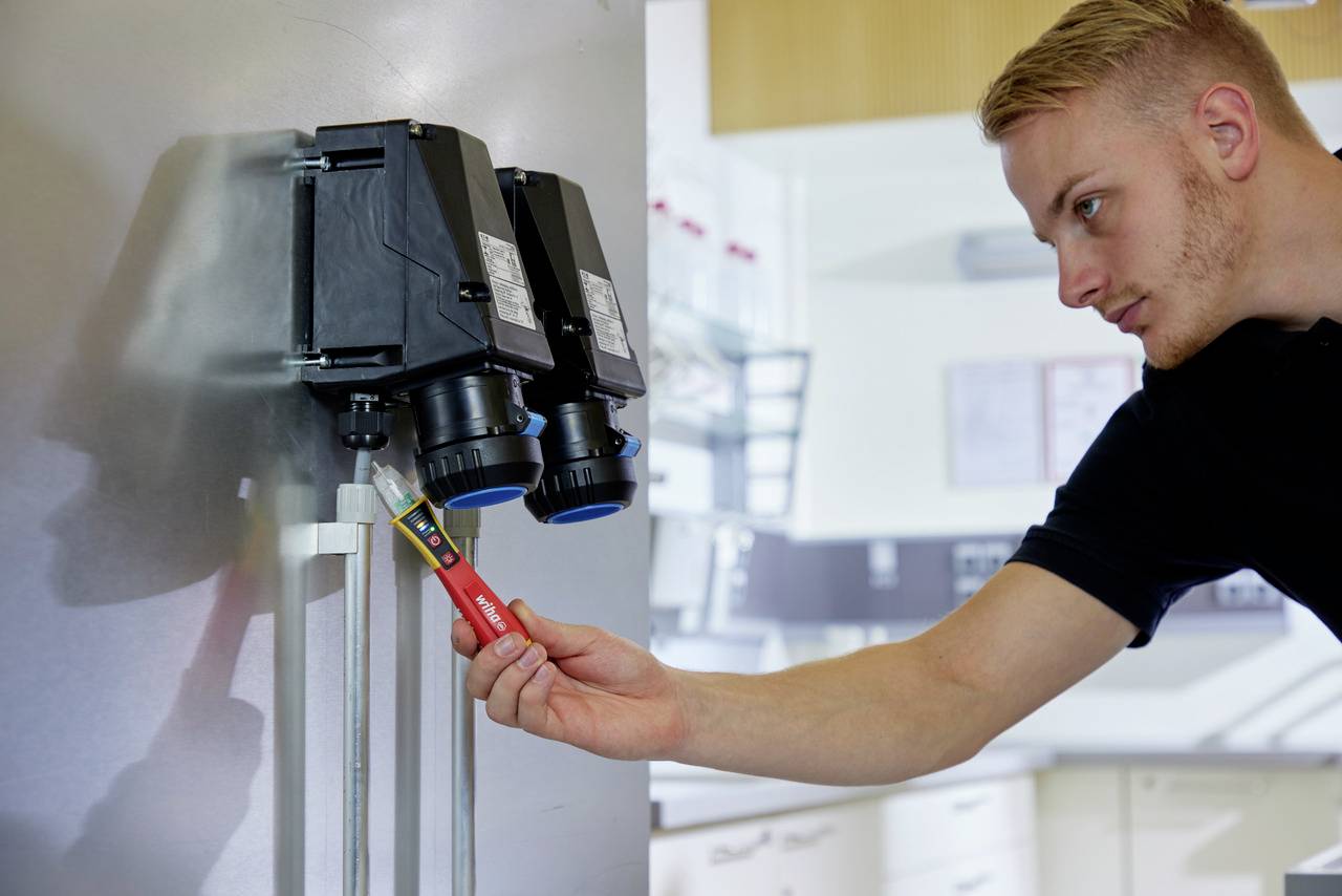Un homme vérifie les connexions électriques dans un armoire électrique à l'aide d'un testeur de tension. L'arrière-plan montre un environnement de laboratoire.