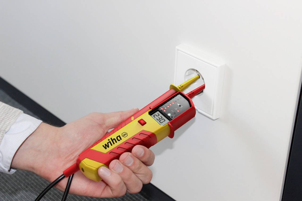 Une personne tient un multimètre numérique contre une prise électrique. L'appareil affiche '230V' sur son écran.