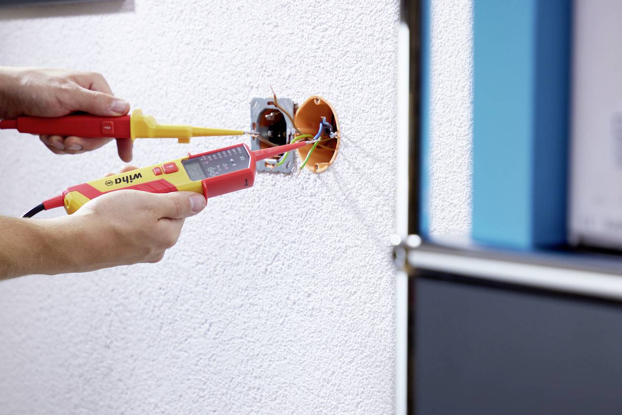 Une personne effectue des mesures avec un multimètre numérique sur des fils électriques dans une prise murale.