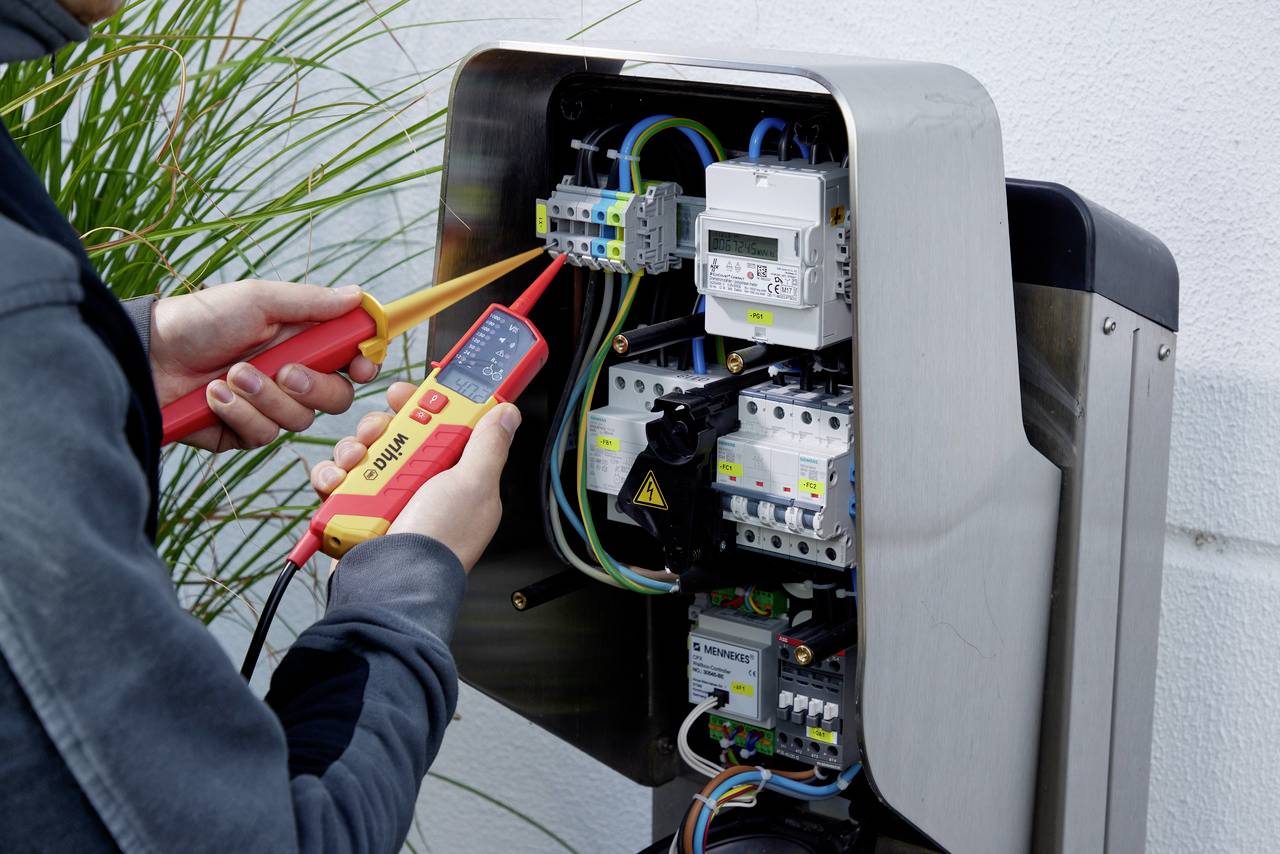 'Une personne teste des connexions électriques dans un armoire électrique ouverte à l'aide d'un multimètre. Des fils et des fusibles sont visibles.'