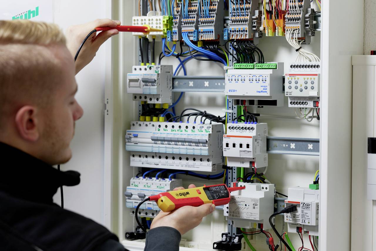 Un technicien vérifie le câblage dans un boîtier électrique ouvert à l'aide d'un appareil de mesure.