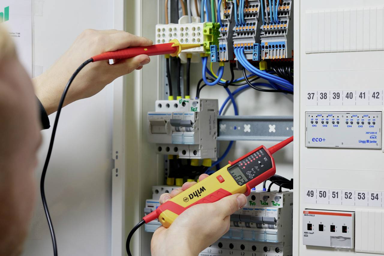 Une personne teste des câbles dans un armoire électrique ouverte à l'aide d'un appareil de mesure électronique. Plusieurs fils de différentes couleurs sont visibles.