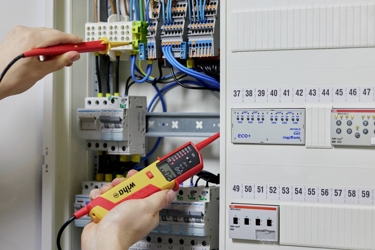 Une personne vérifie les connexions électriques à l'aide d'un multimètre dans un armoire électrique ouverte remplie de câbles et de fusibles.