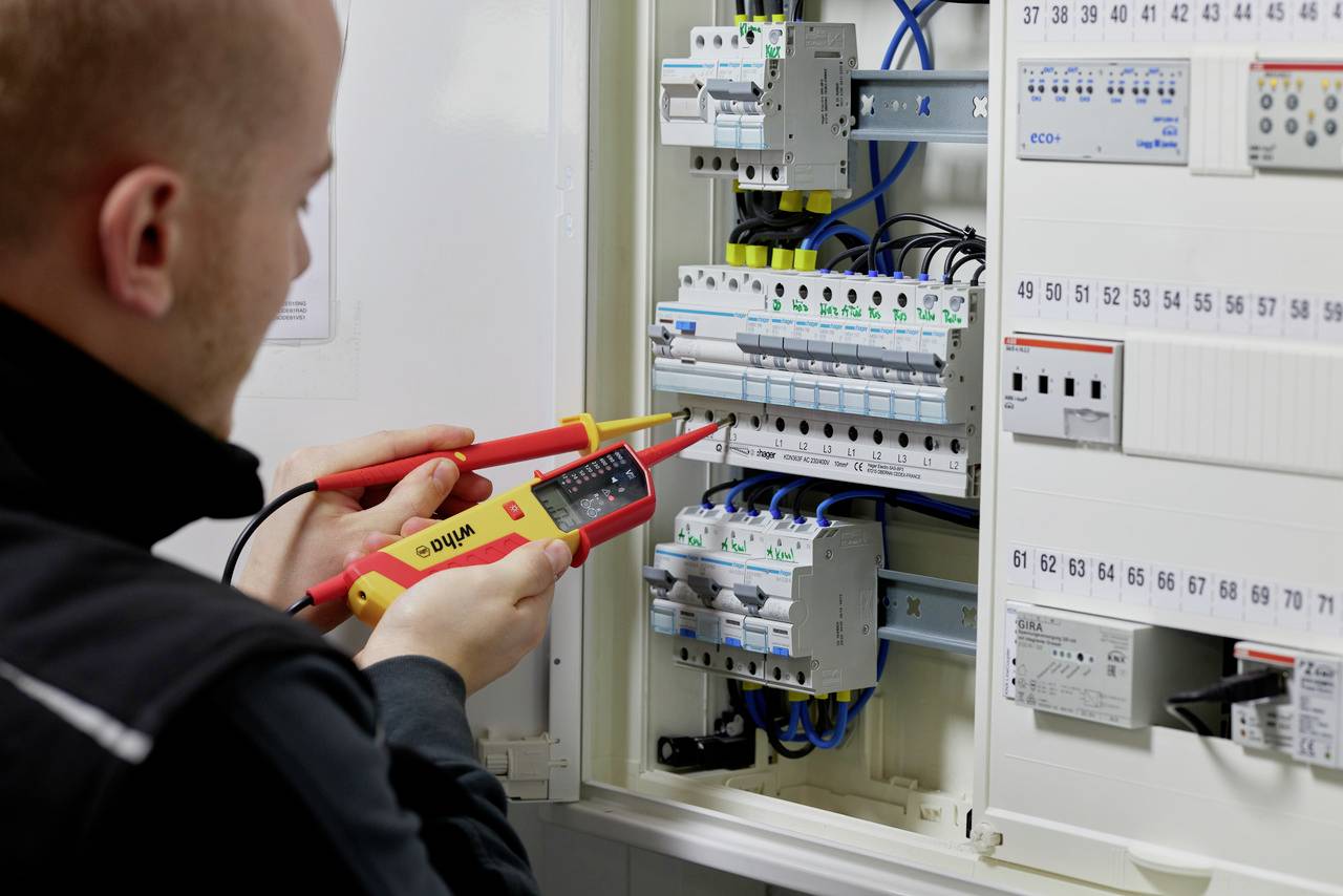 Une personne vérifie les connexions électriques dans un tableau électrique ouvert à l'aide d'un multimètre.