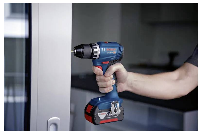 Une personne tient une perceuse sans fil bleue et noire près d'une surface métallique à l'intérieur d'une pièce moderne, suggérant un travail de bricolage ou d'amélioration domiciliaire.