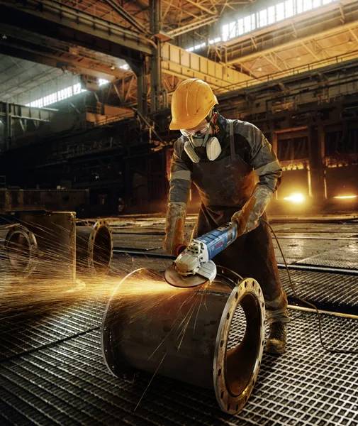 Un travailleur, portant un casque de protection et un masque, utilise un outil de meulage sur un grand tuyau métallique dans un environnement industriel d'usine.