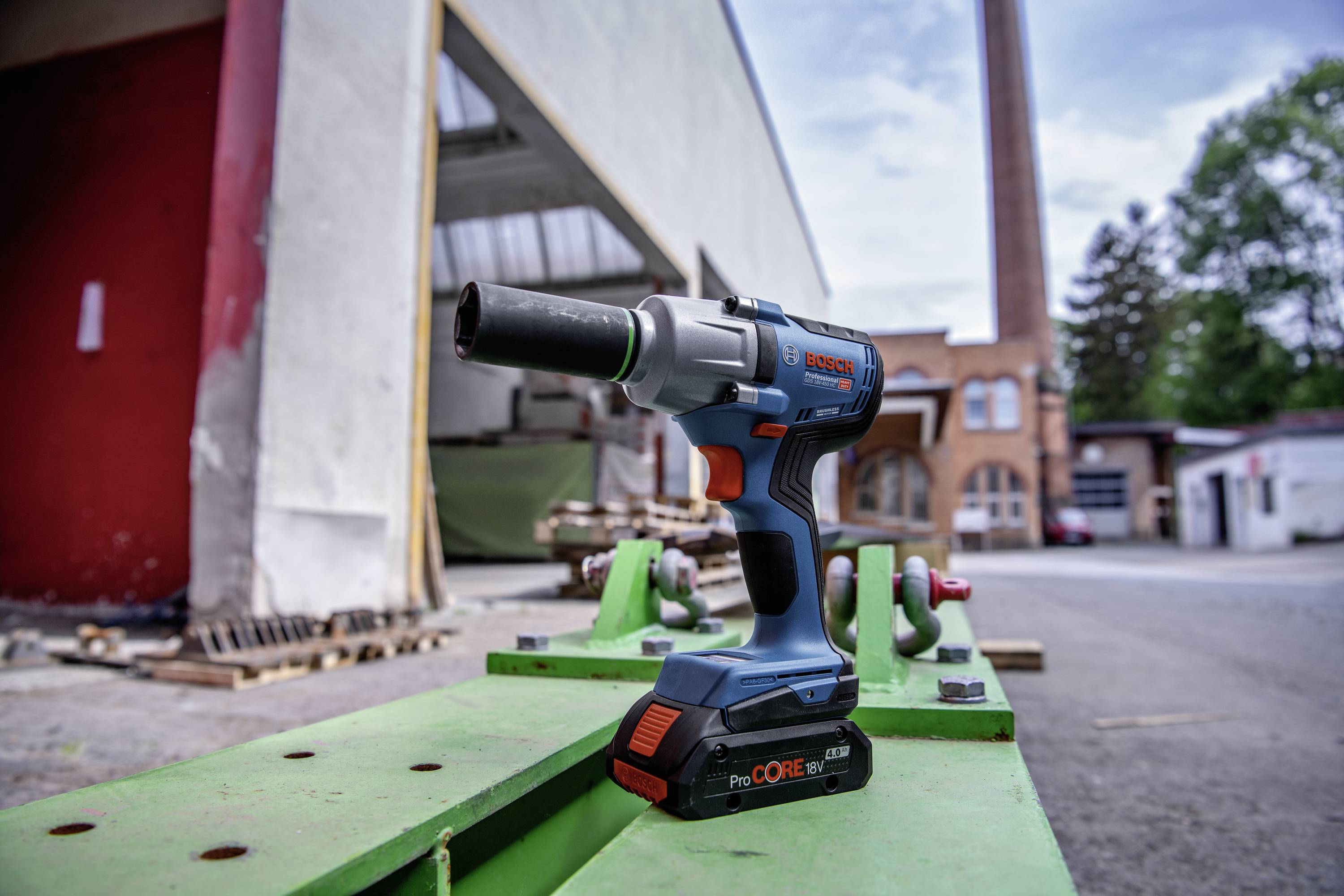 Un tournevis sans fil bleu et orange est posé sur une table verte, avec en arrière-plan un hall d'usine et une cheminée.