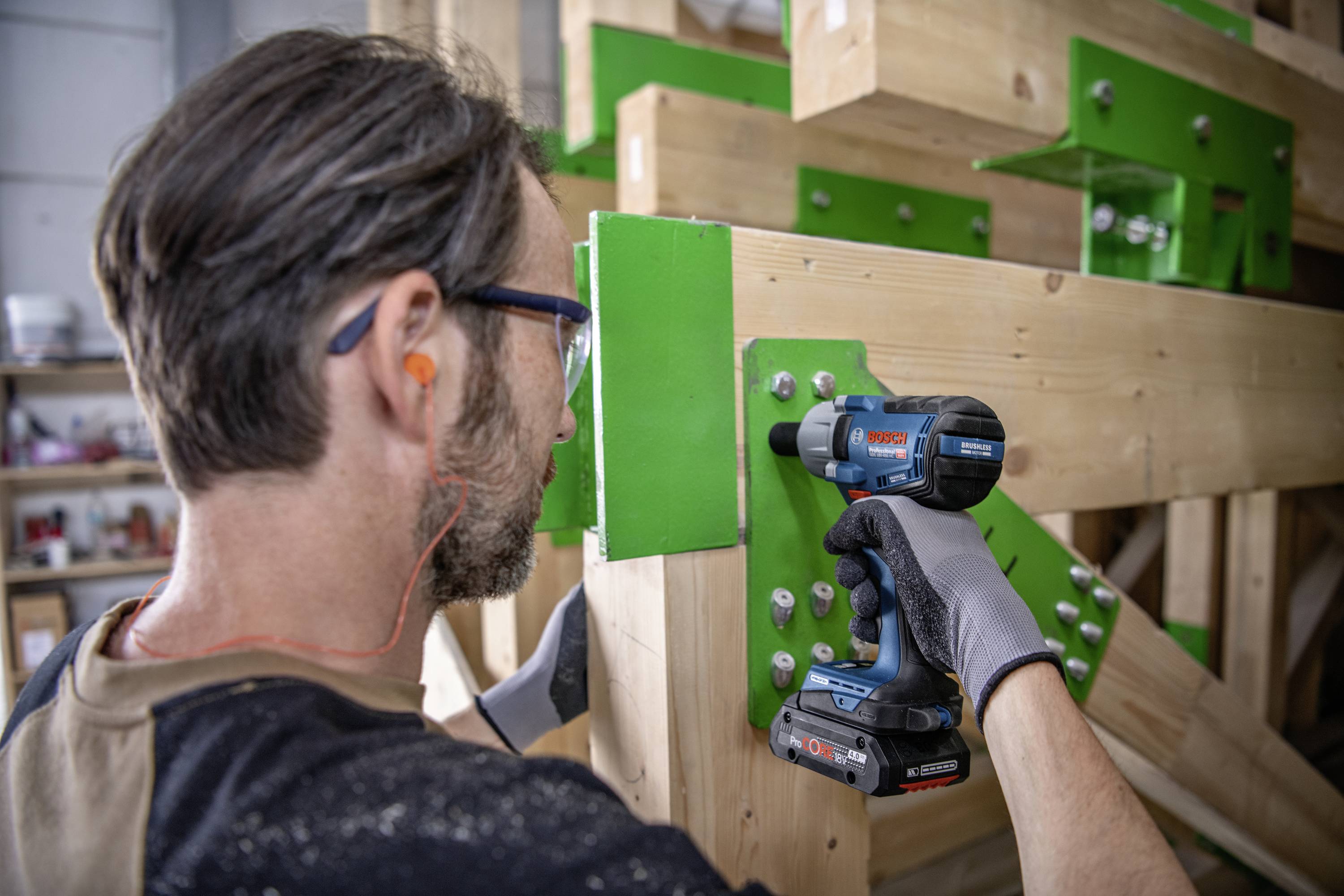 Un artisan fixe des poutres en bois avec une visseuse sans fil. Il porte des protections auditives et des gants de travail, et travaille sur une construction en bois.